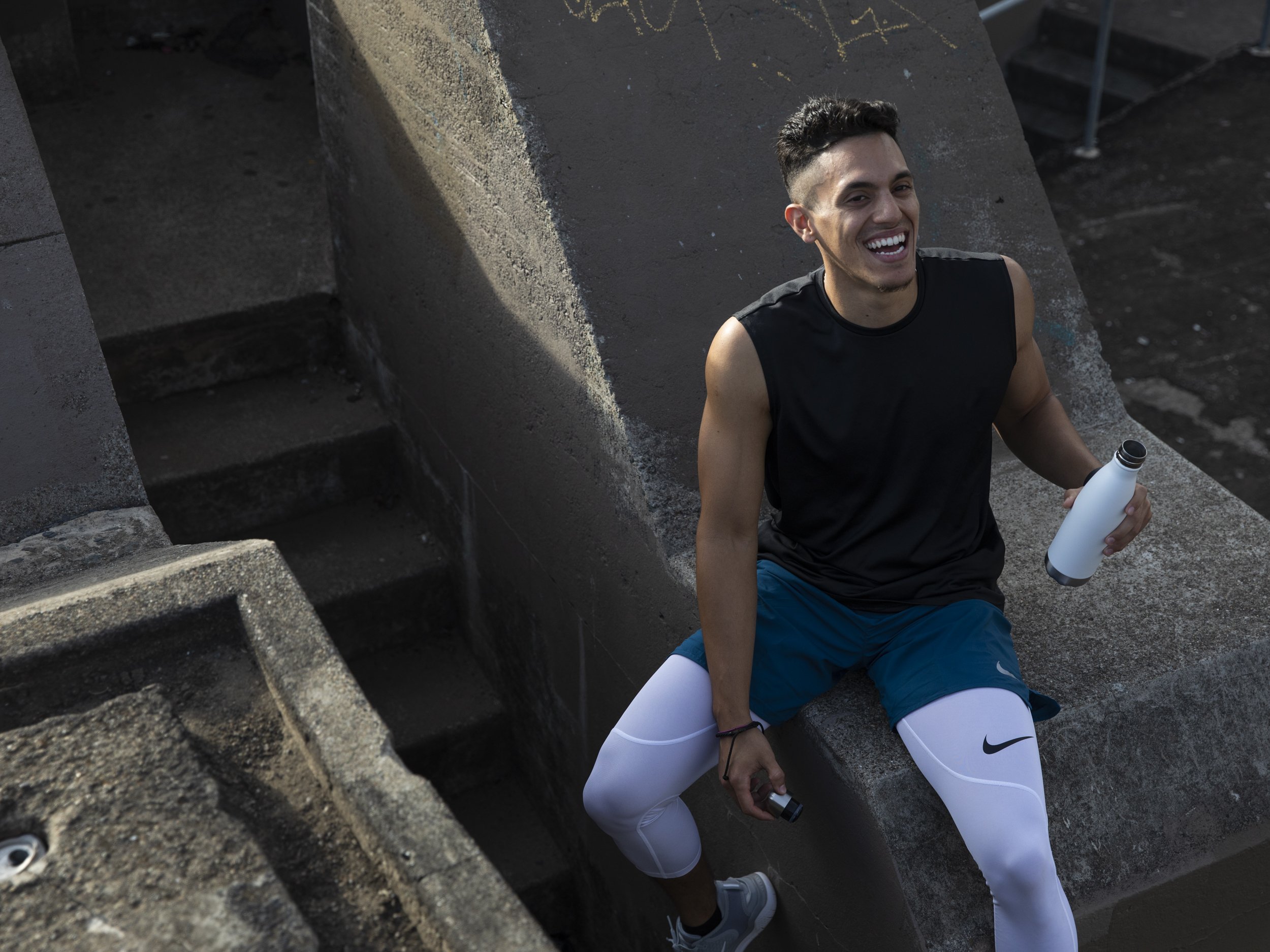 Athletic fitness and training photography by Bay Area commercial photographer Kaare Iverson for Future Fit App. A young man with short dark hair, wearing Nike leggings, is sitting on concrete stairs outdoors, smiling and holding a white water bottle.