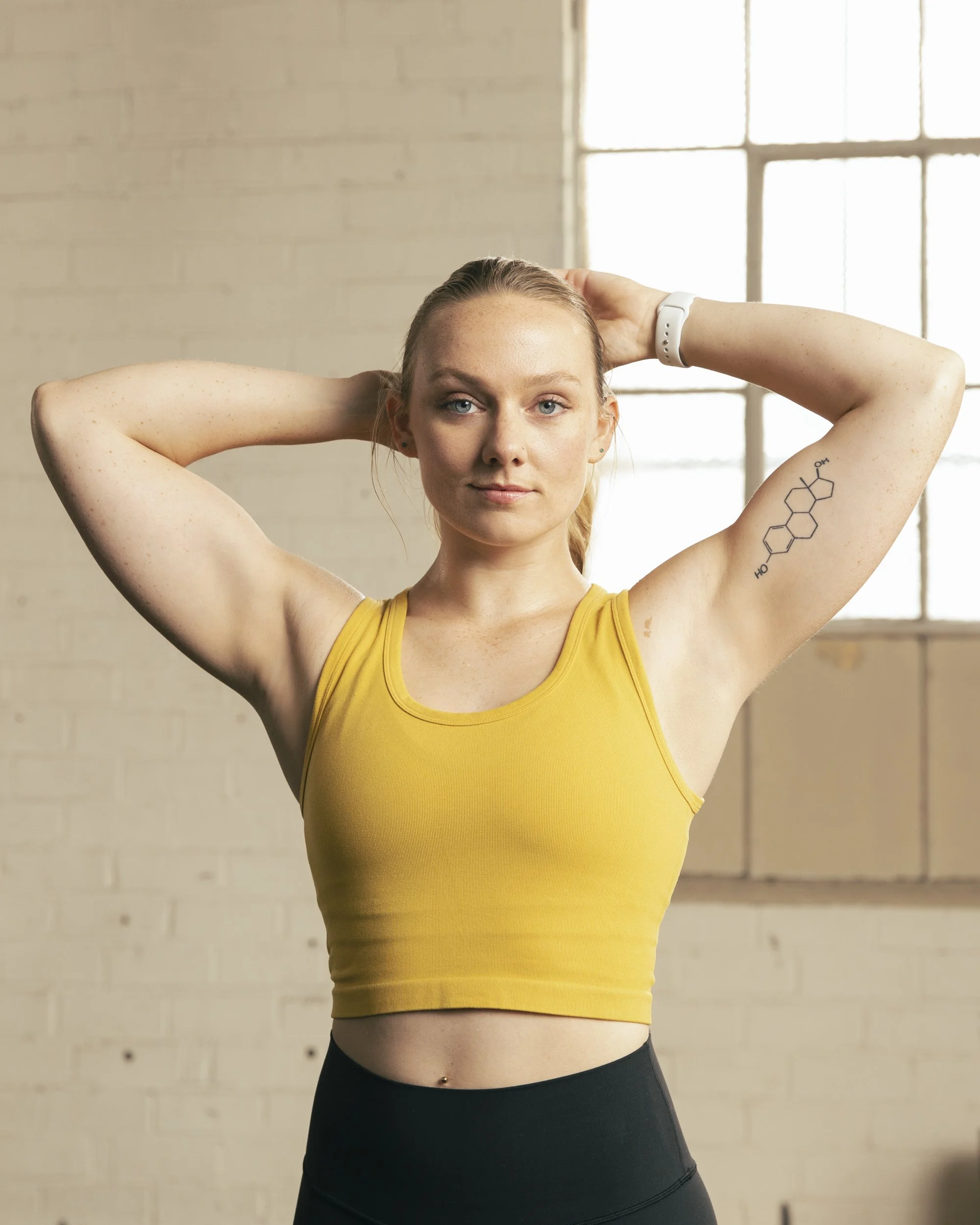 Athletic fitness photography by Bay Area commercial photographer Kaare Iverson. A young woman with blonde hair, wearing a yellow sports bra, and a white fitness tracker, standing with arms raised behind her head in a crossfit gym.Shot for Future App.
