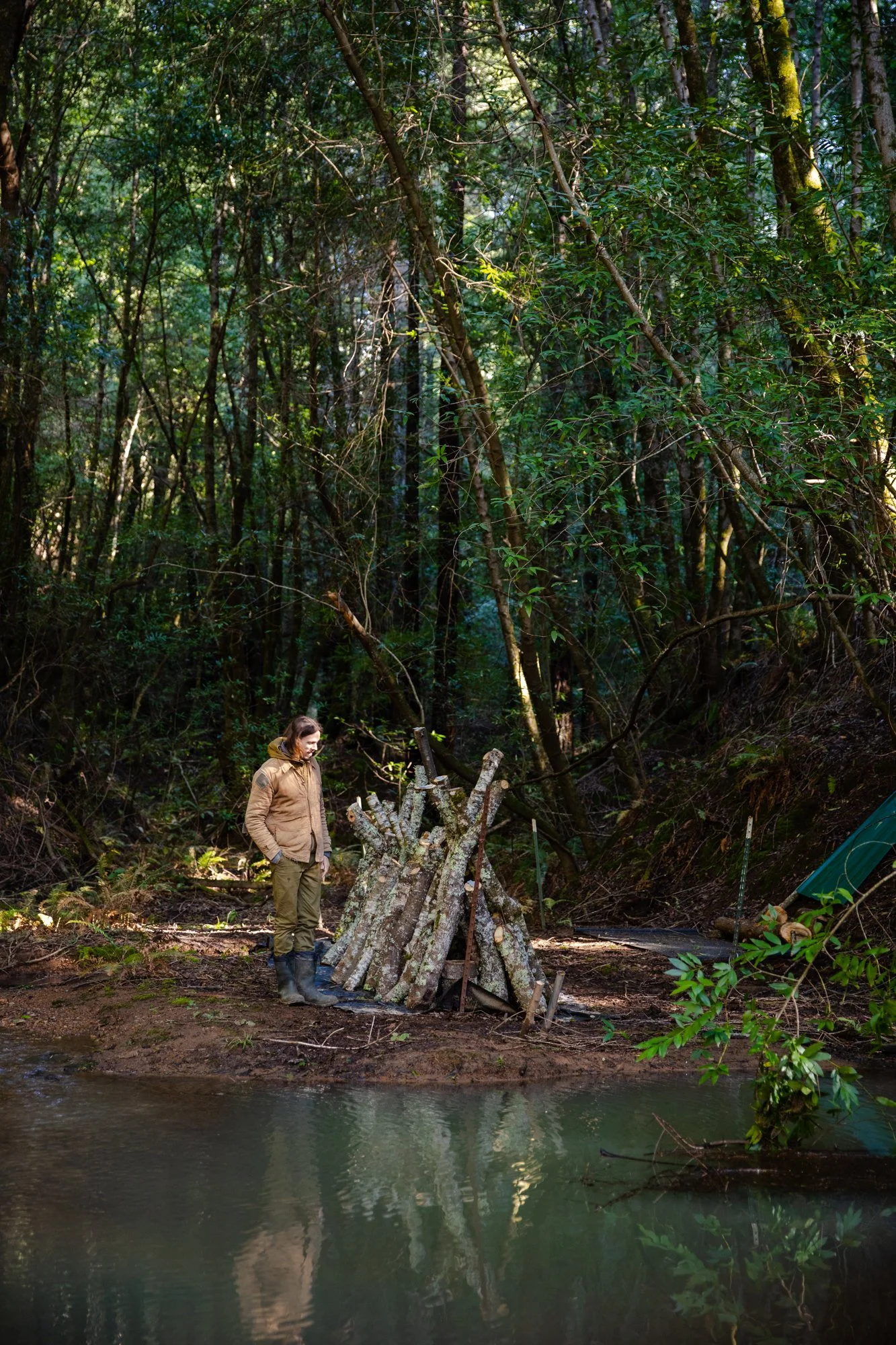 Image from PRAXIS, a photo essay shot in California by photographer Kaare Iverson. A person standing by a stack of cut logs for mushroom growing near a river in a dense forest.