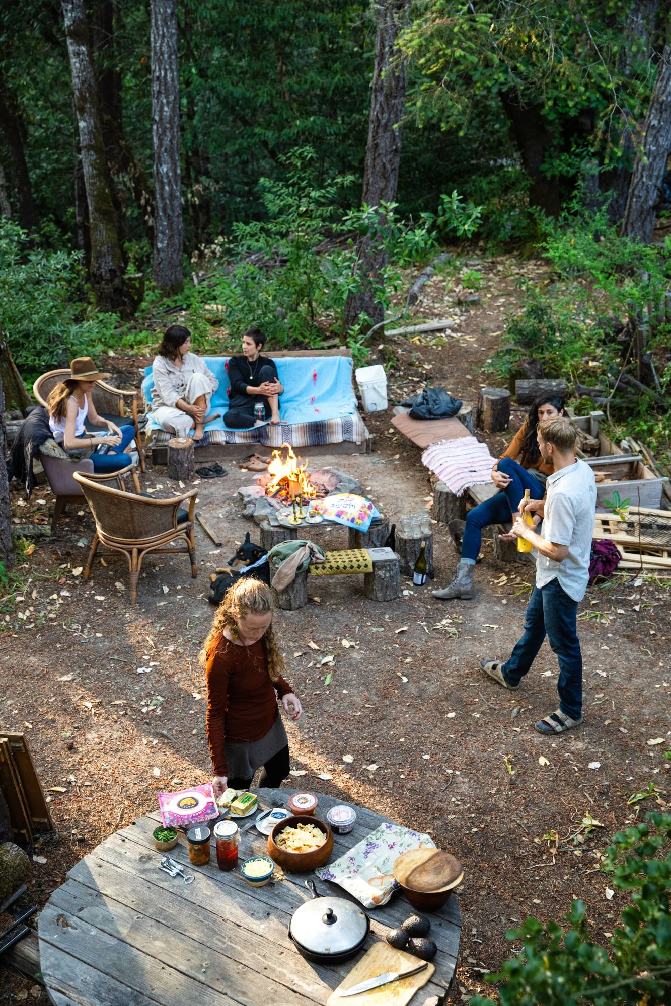 Image from PRAXIS, a photo essay shot in California by photographer Kaare Iverson. Friends gathering around a campfire in a forest, sitting on sofas and chairs, with food and drinks on tables.