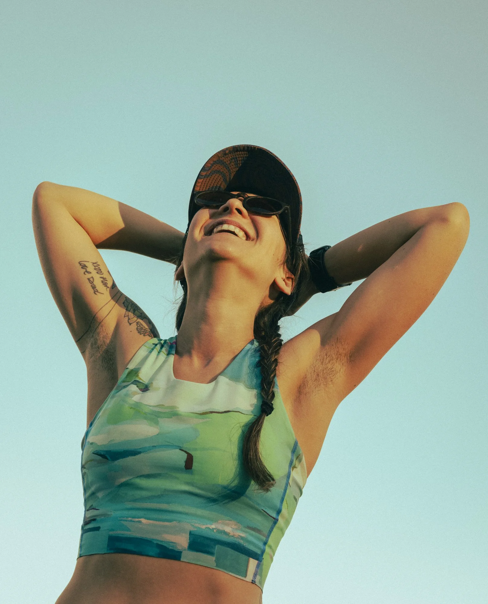 Athletic wear fitness lifestyle photography by San Francisco based photographer Kaare Iverson. A woman in a colorful Janji tank top and a cap with sunglasses, smiling with her arms behind her head, against a clear blue sky.