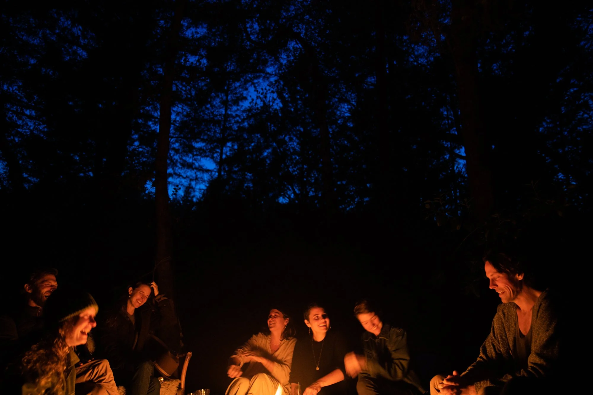 Image from PRAXIS, a photo essay shot in California by photographer Kaare Iverson. A group of people sitting around a campfire at night in a forest, smiling and laughing with tall trees and a dark blue sky in the background.