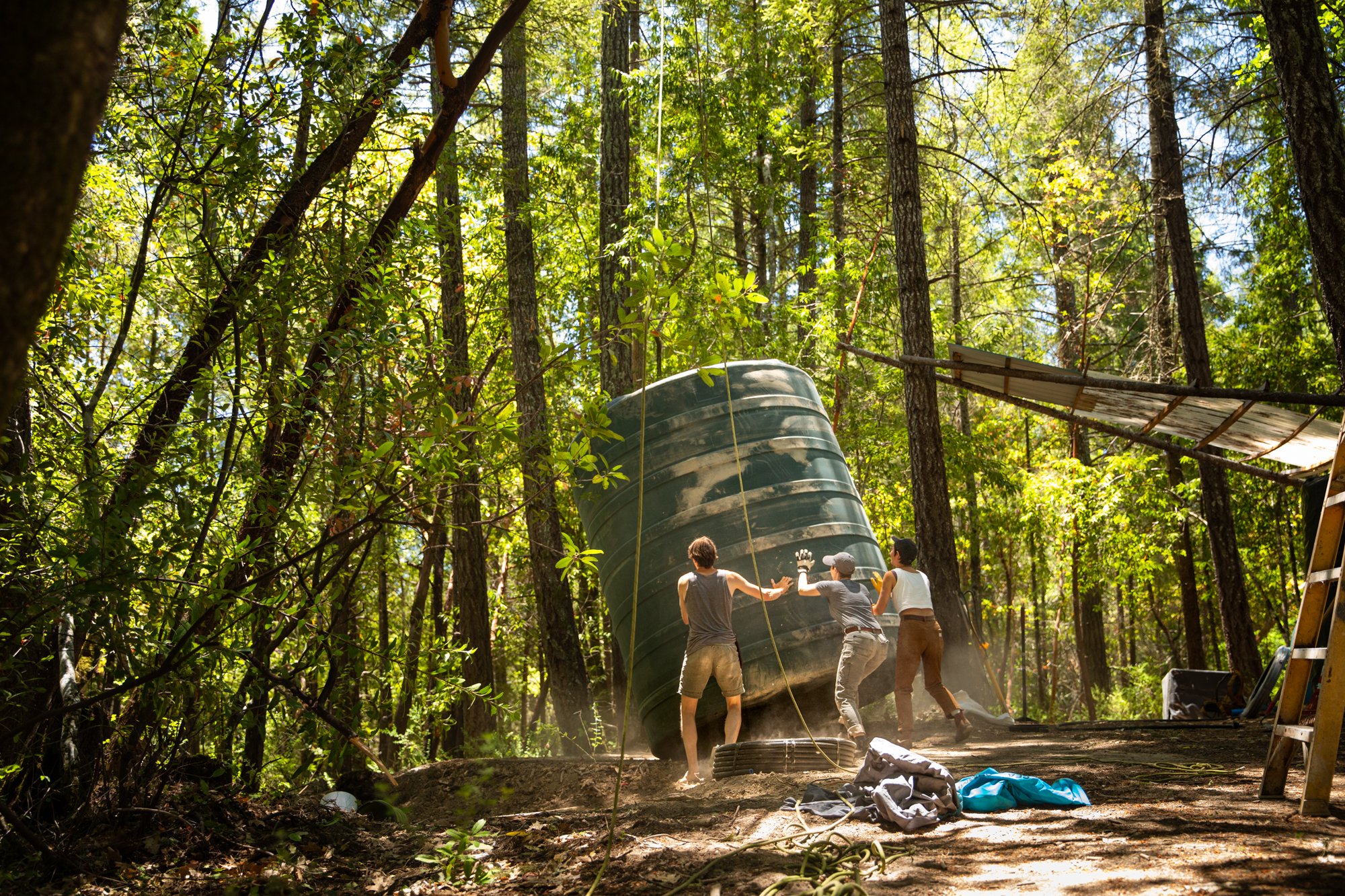 Image from the photo essay PRAXIS about alternative living in California by photographer, Kaare Iverson. Group of people working outdoors in a wooded area, installing a large green plastic water tank, with sunlight filtering through the trees.