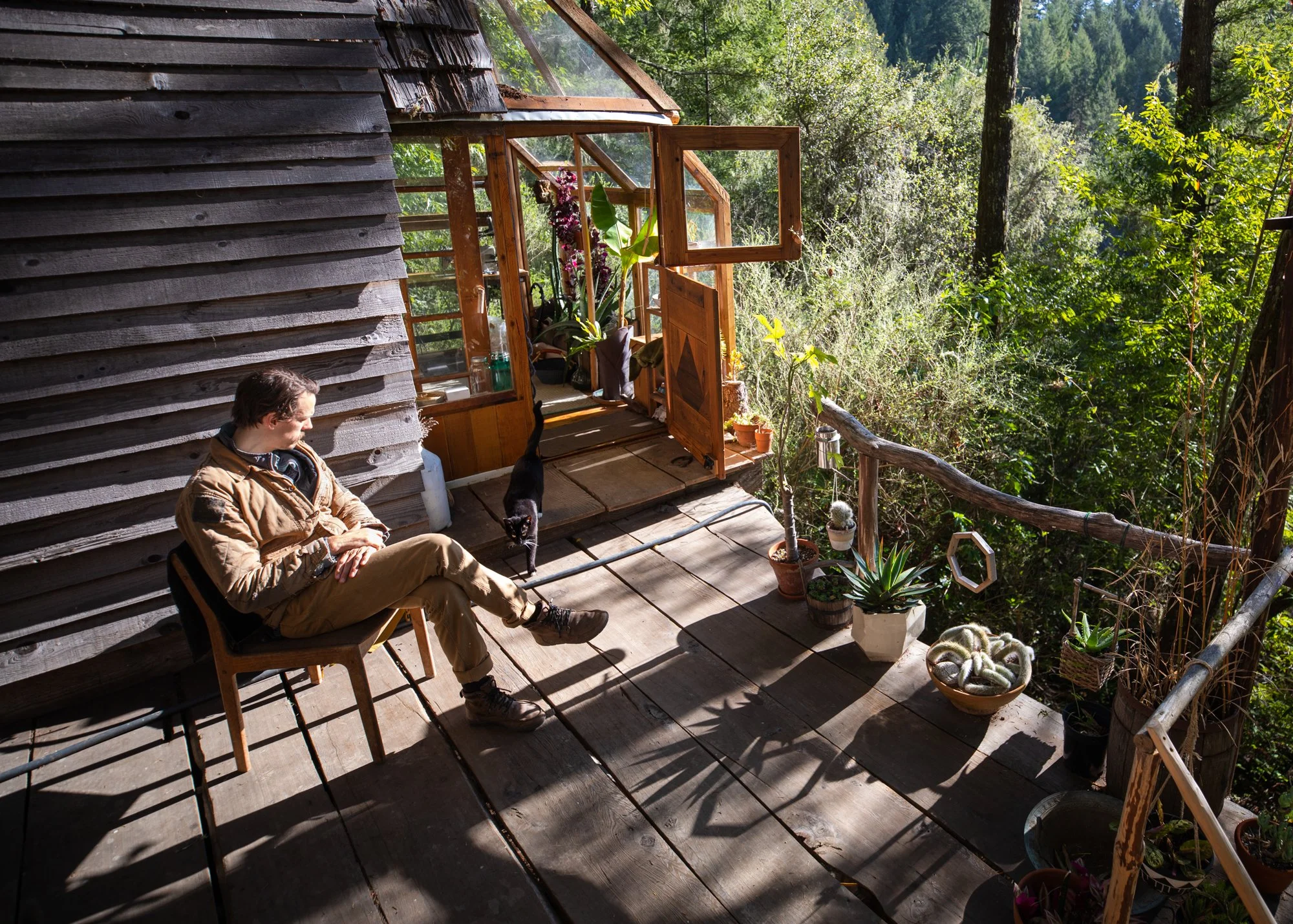 Image from the photo essay PRAXIS about alternative living in California by photographer, Kaare Iverson. A man sitting on a chair on a wooden balcony in a forested area, with potted plants and a black cat nearby.