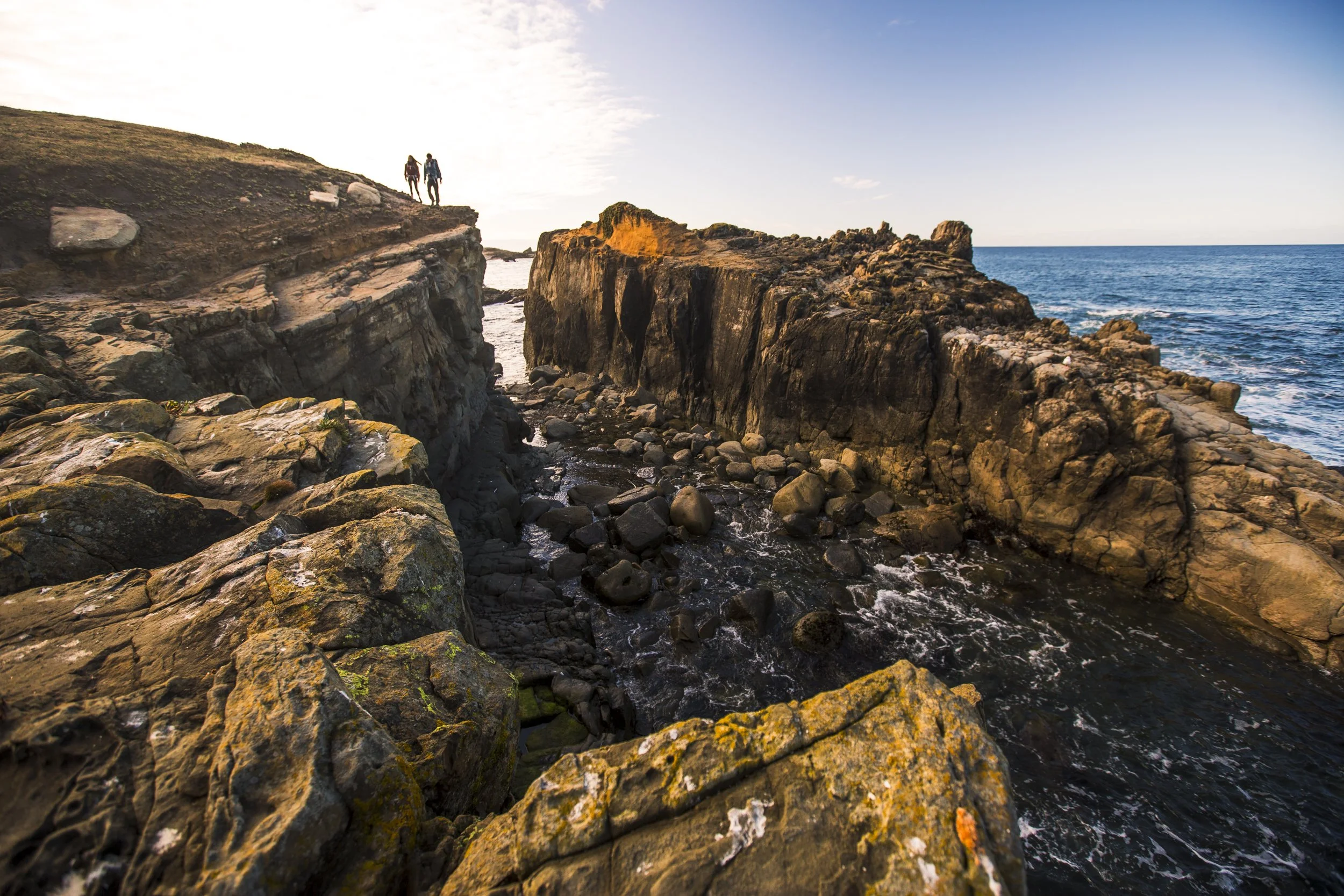 Epic landscape lifestyle photography shot for Osprey Pack product launch in Sonoma County, California by Kaare Iverson. Two people walking along a rocky coastal cliff at sunset, overlooking the ocean.
