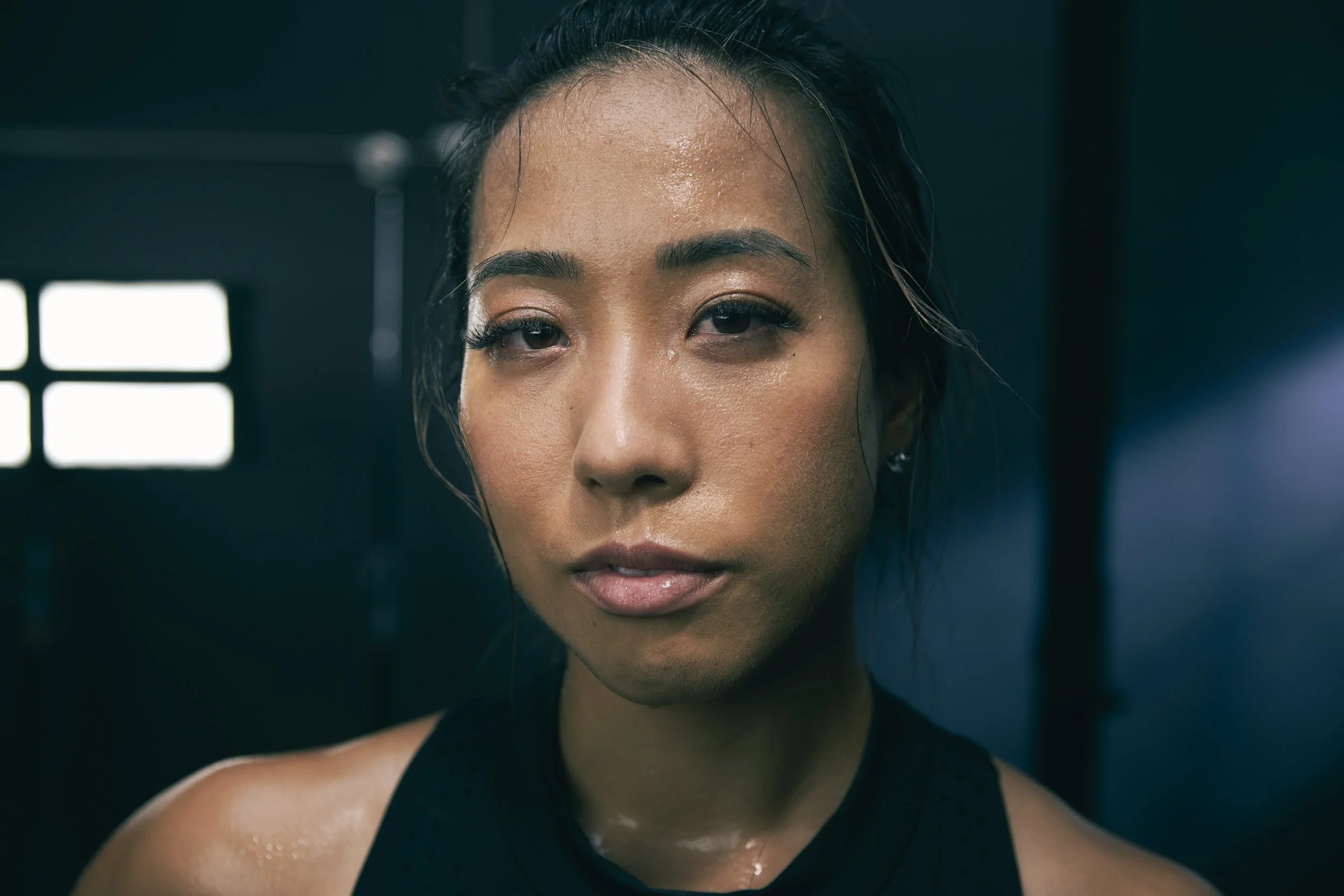 Fitness lifestyle advertising image shot for Bay Area technology company Future Fit by commercial photographer Kaare Iverson. Close-up of a woman with glowing skin and dark hair in a gym environment with dim, dramatic lighting.