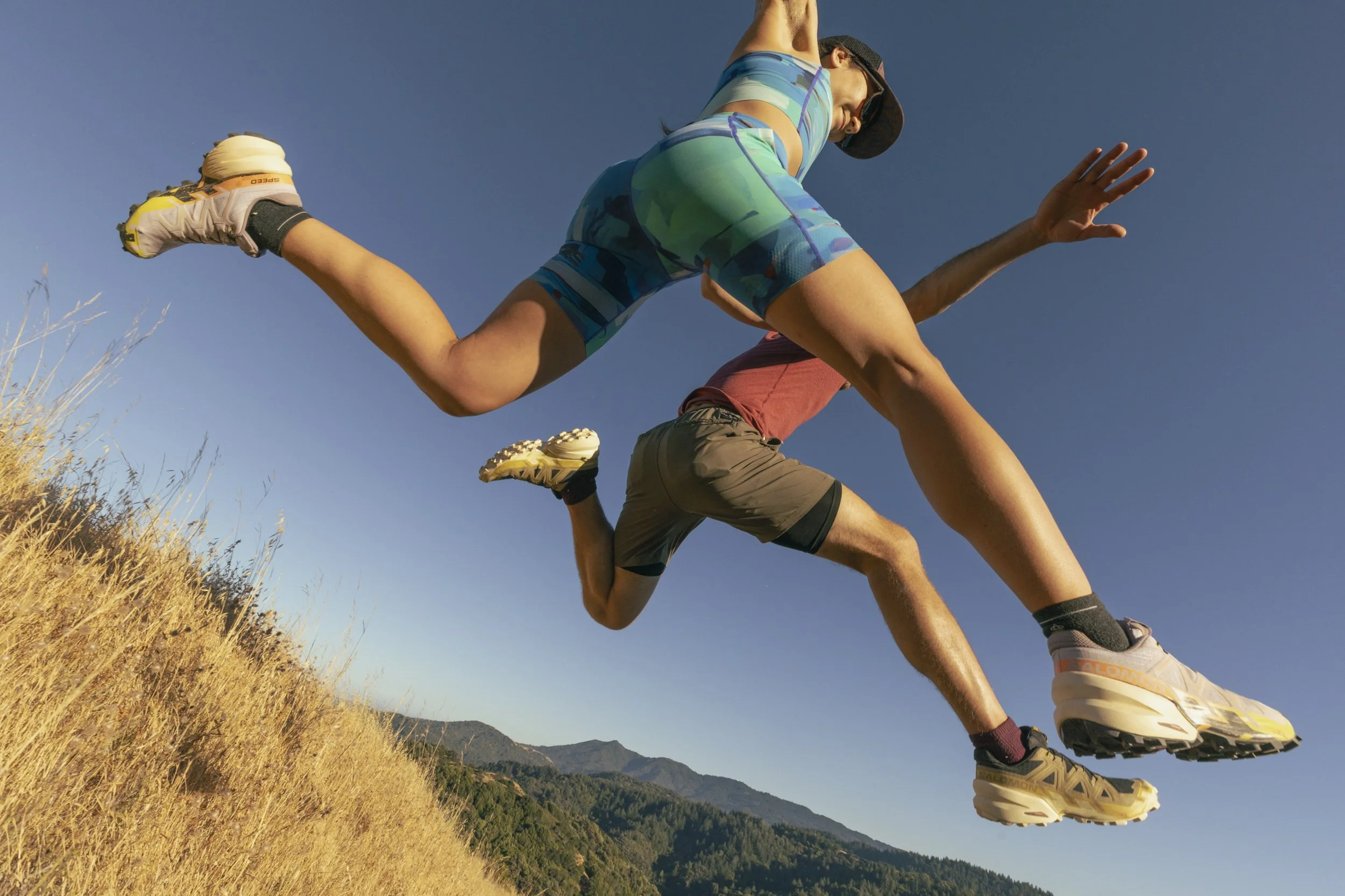 Athletic wear fitness lifestyle photography by San Francisco based photographer Kaare Iverson. Two trail runners in Salomon shoes leap over camera on a sunny day.