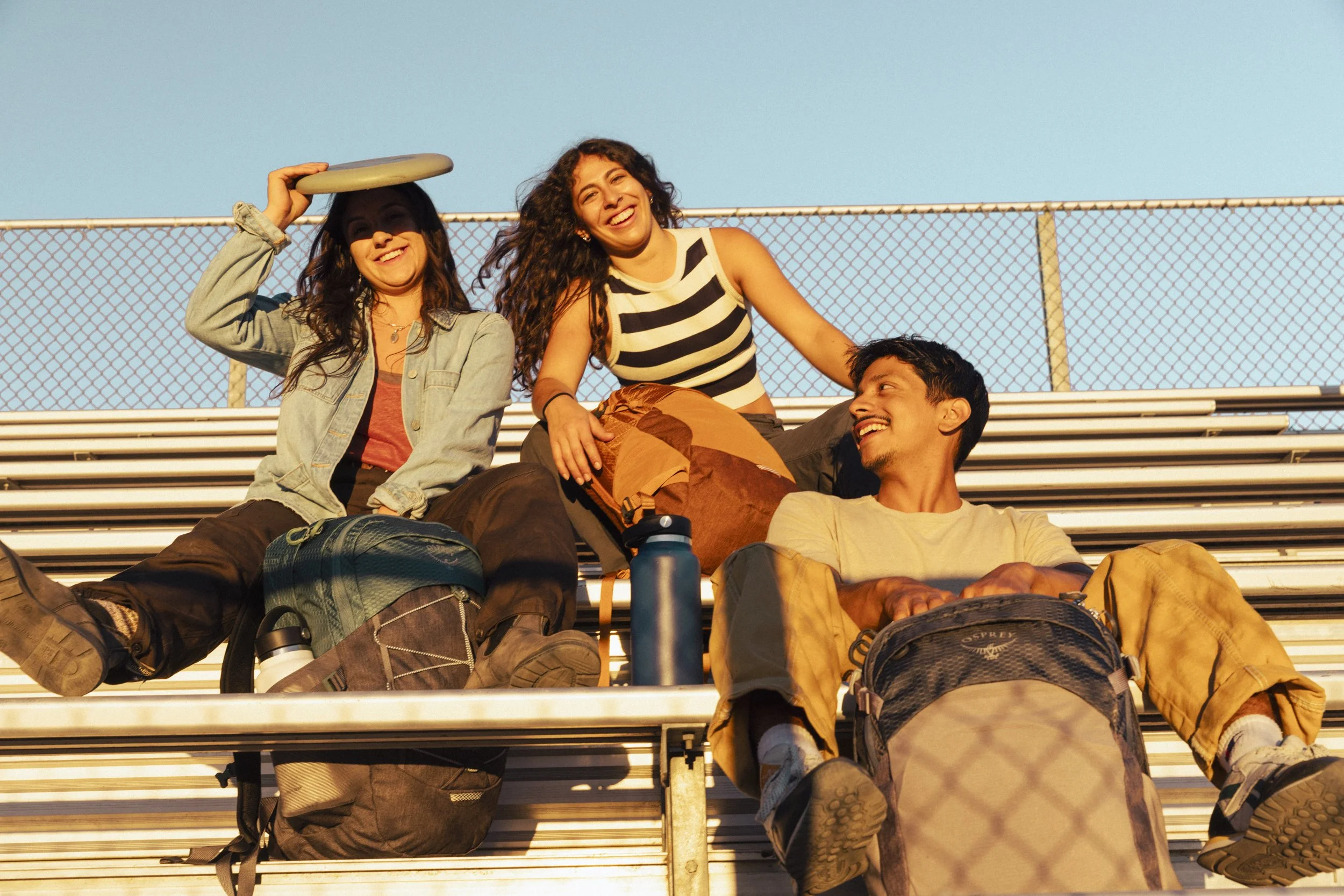 College lifestyle photography by commercial photographer Kaare Iverson for Osprey Packs. Three friends sitting on metal bleachers outdoors, smiling and enjoying each other's company during sunset. Shot in California.