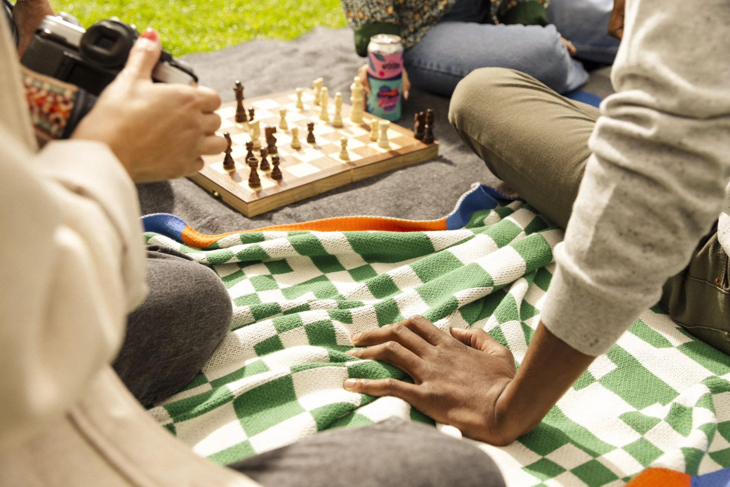 Urban park lifestyle image by commercial photographer Kaare Iverson for Treasure Island Development Group.  Close-up of people playing chess outdoors, sitting on a blanket on the grass, with drinks nearby.