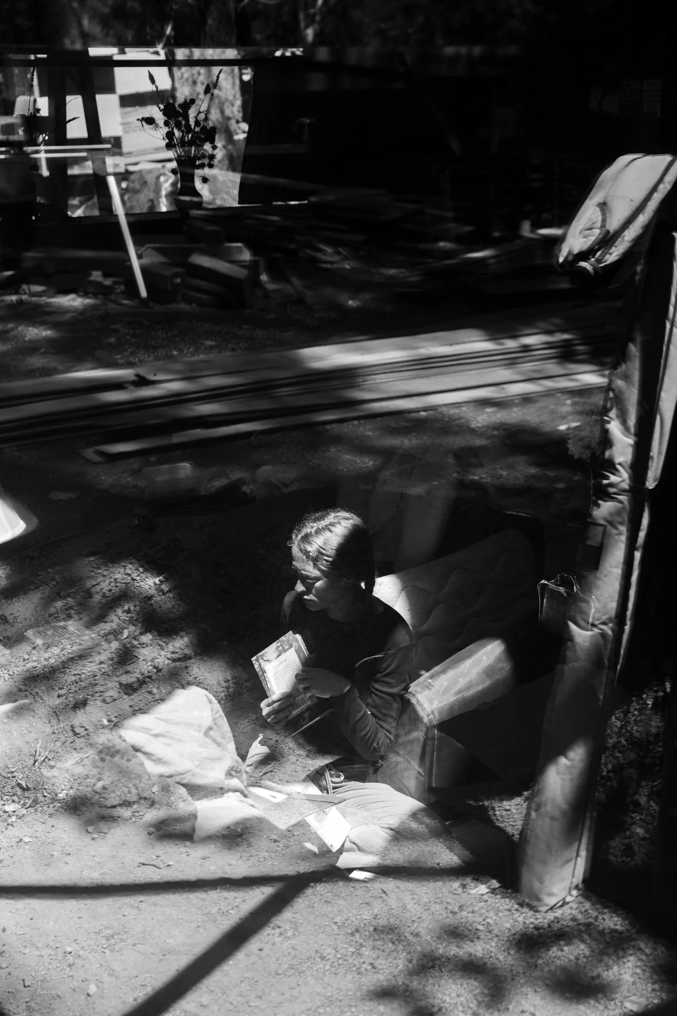 Image from the photo essay PRAXIS about alternative living in California by photographer, Kaare Iverson. A black and white photo taken through a glass window showing a woman packing her belongings. Shadows and reflections are visible on the glass.