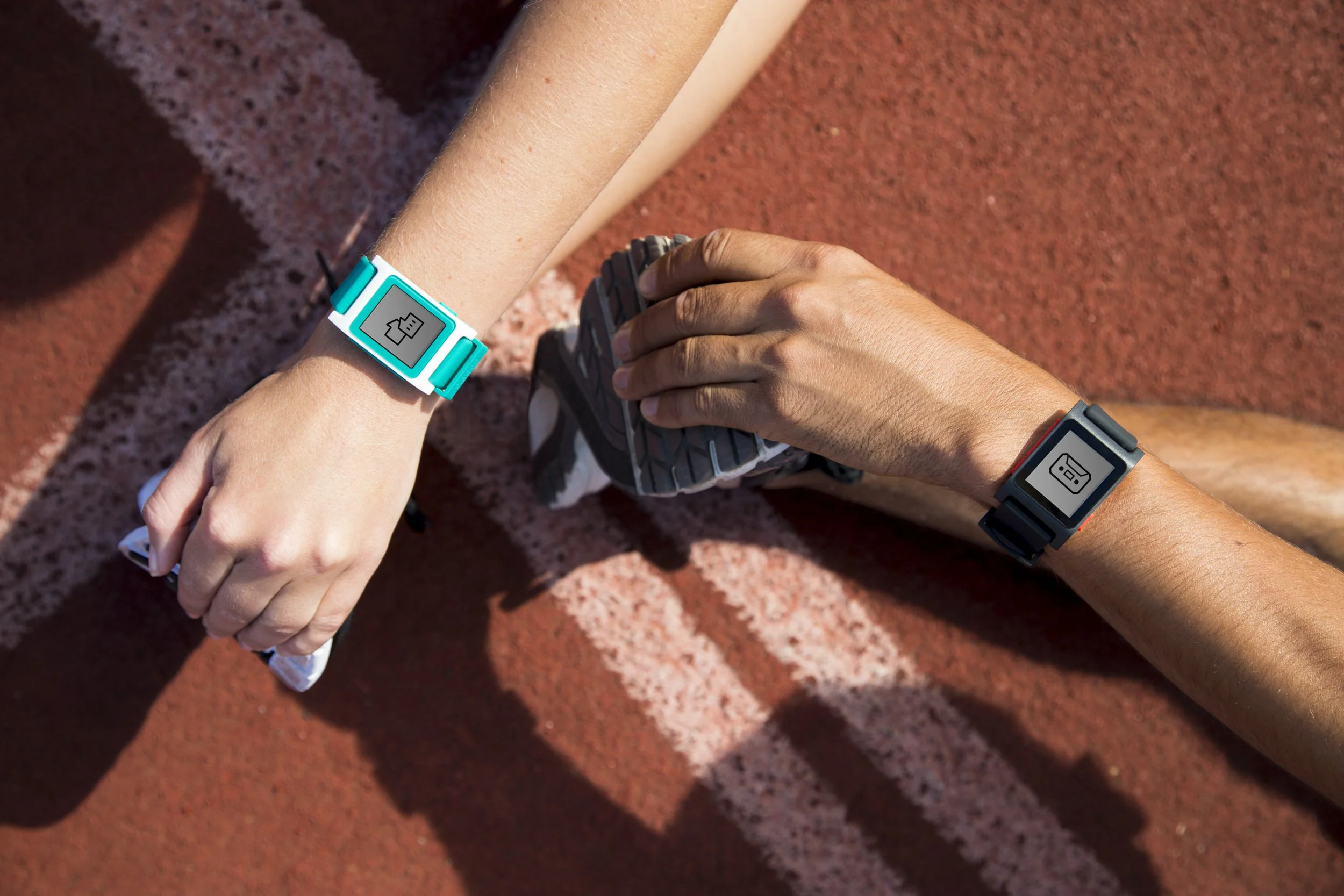 Close-up of two athletes wearing digital fitness watches on their wrists, one with a blue band and the other with a dark band, on a red running track.