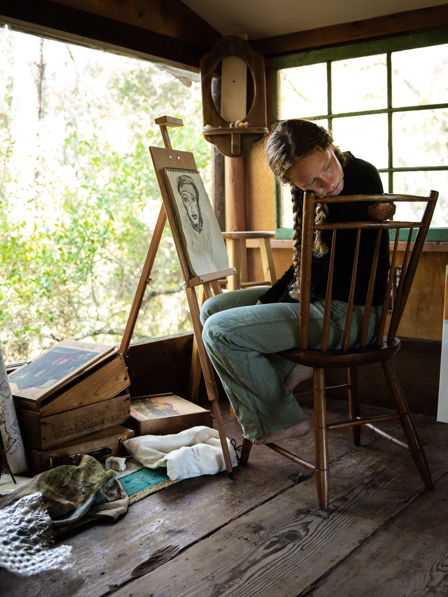 Image from the photo essay PRAXIS about alternative living in California by photographer, Kaare Iverson. Portrait of a sad woman sitting in a wooden chair. There is an easel with a portrait of a woman and sunlight coming through the windows.