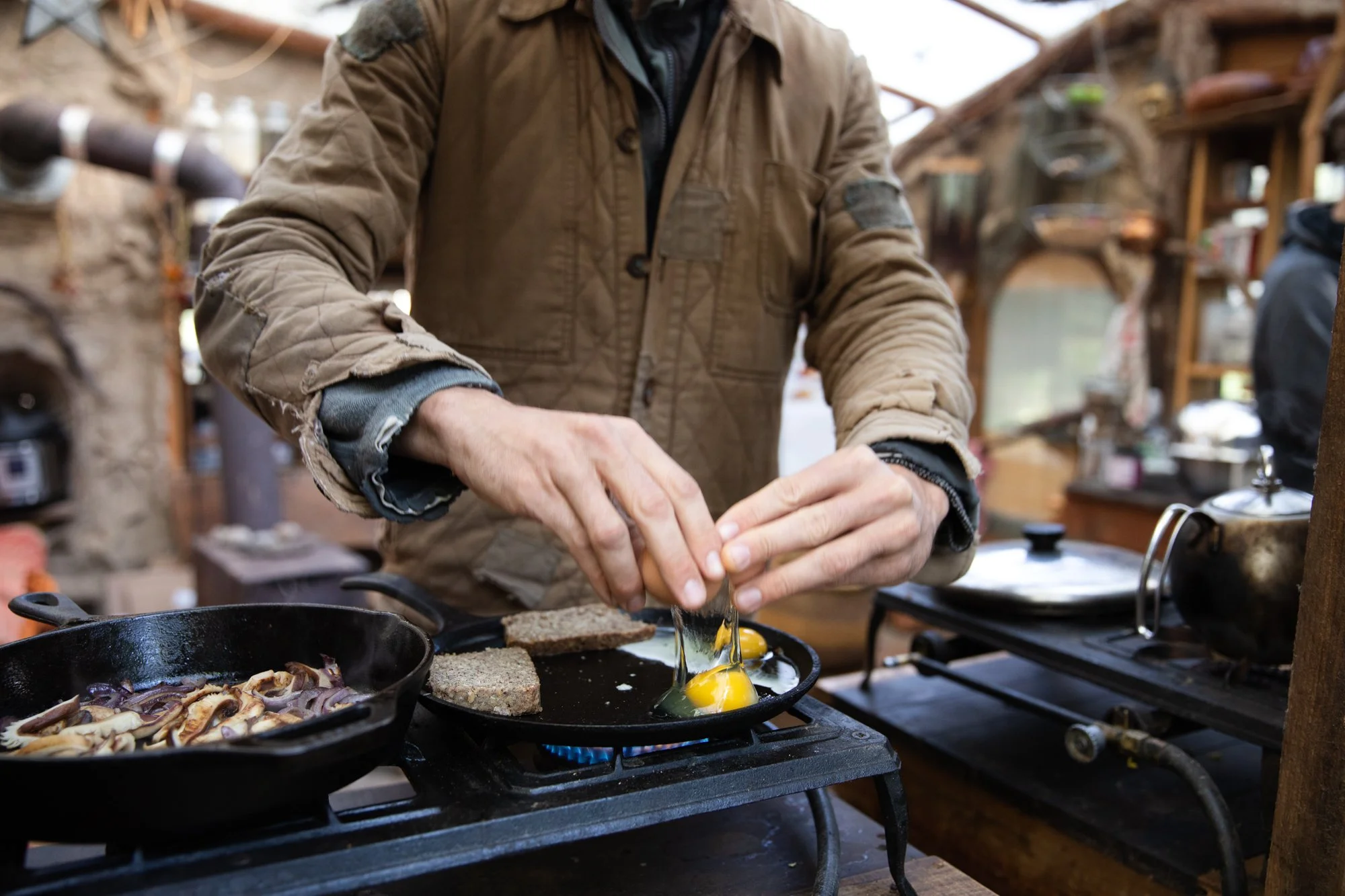Image from the photo essay PRAXIS about alternative living in California by photographer, Kaare Iverson. Person cooking breakfast with a cast iron skillet, eggs in a frying pan, and toast on a stove in a rustic kitchen.