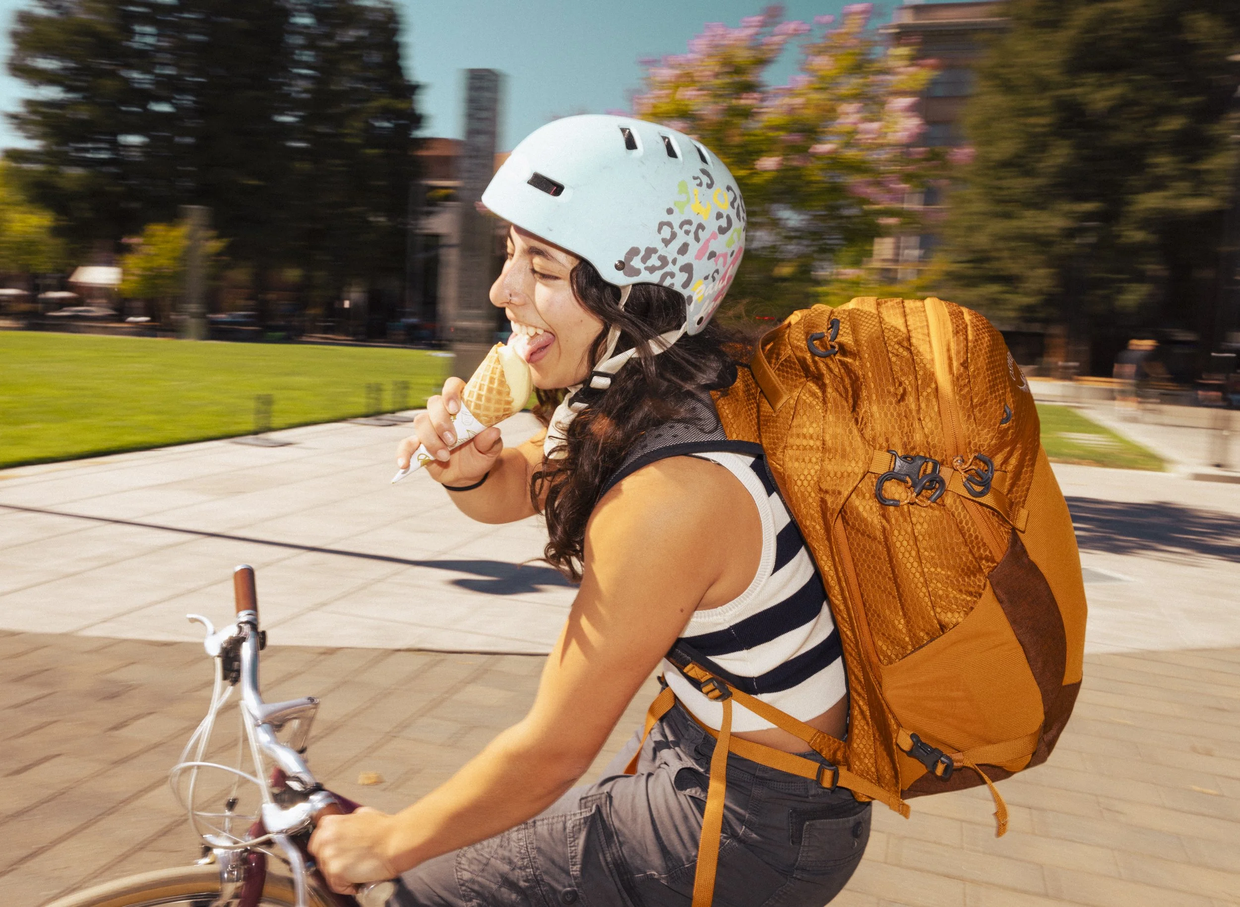 Commercial photography for Osprey Packs of young woman riding a bicycle outdoors, wearing a blue helmet with colorful patterns, a striped shirt, and carrying an orange backpack, smiling and enjoying sunny weather - shot in California.