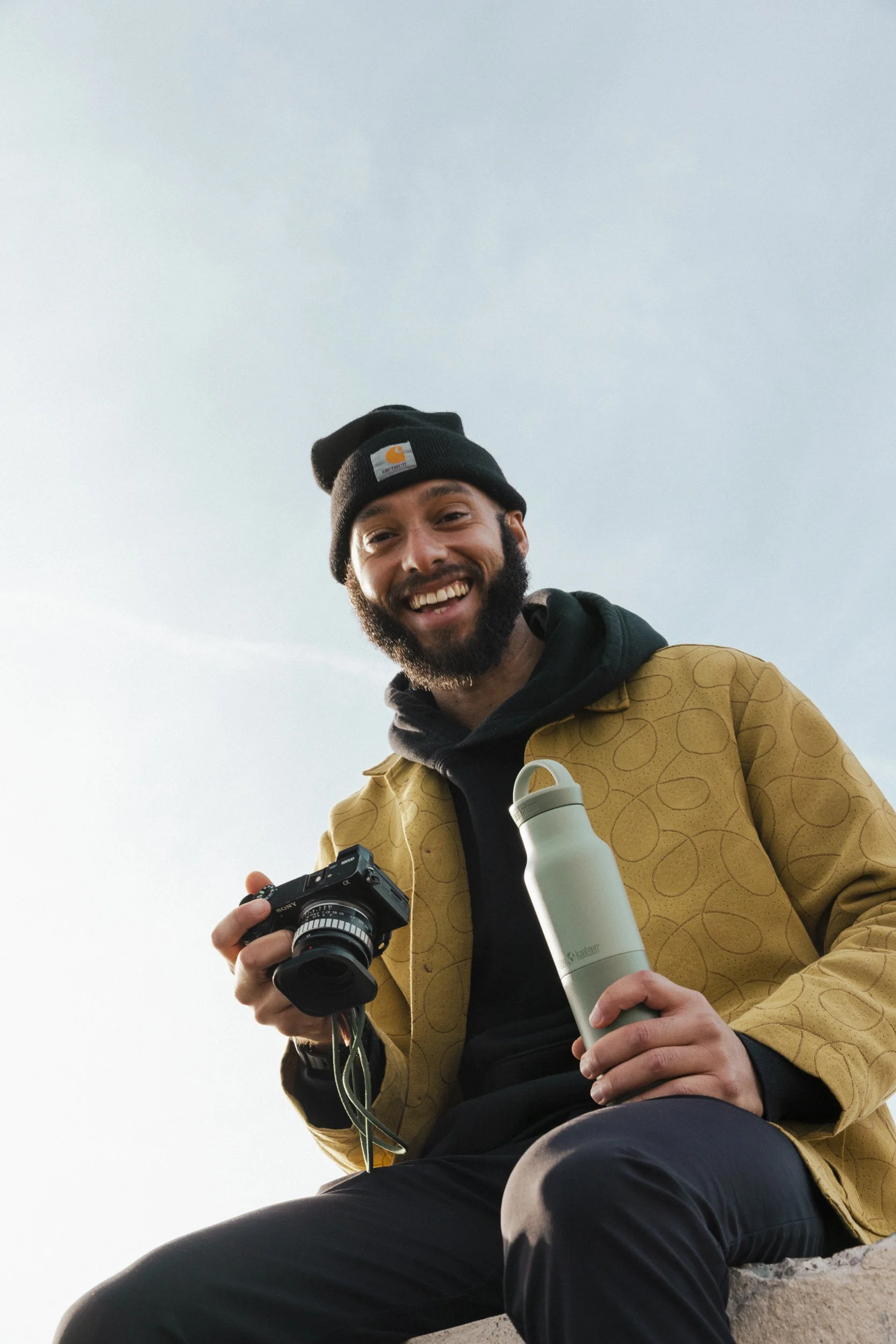 Urban playful lifestyle product photography by Bay Area based photographer Kaare Iverson. man smiles at camera while holding digital camera and Klean Kanteen water bottle.
