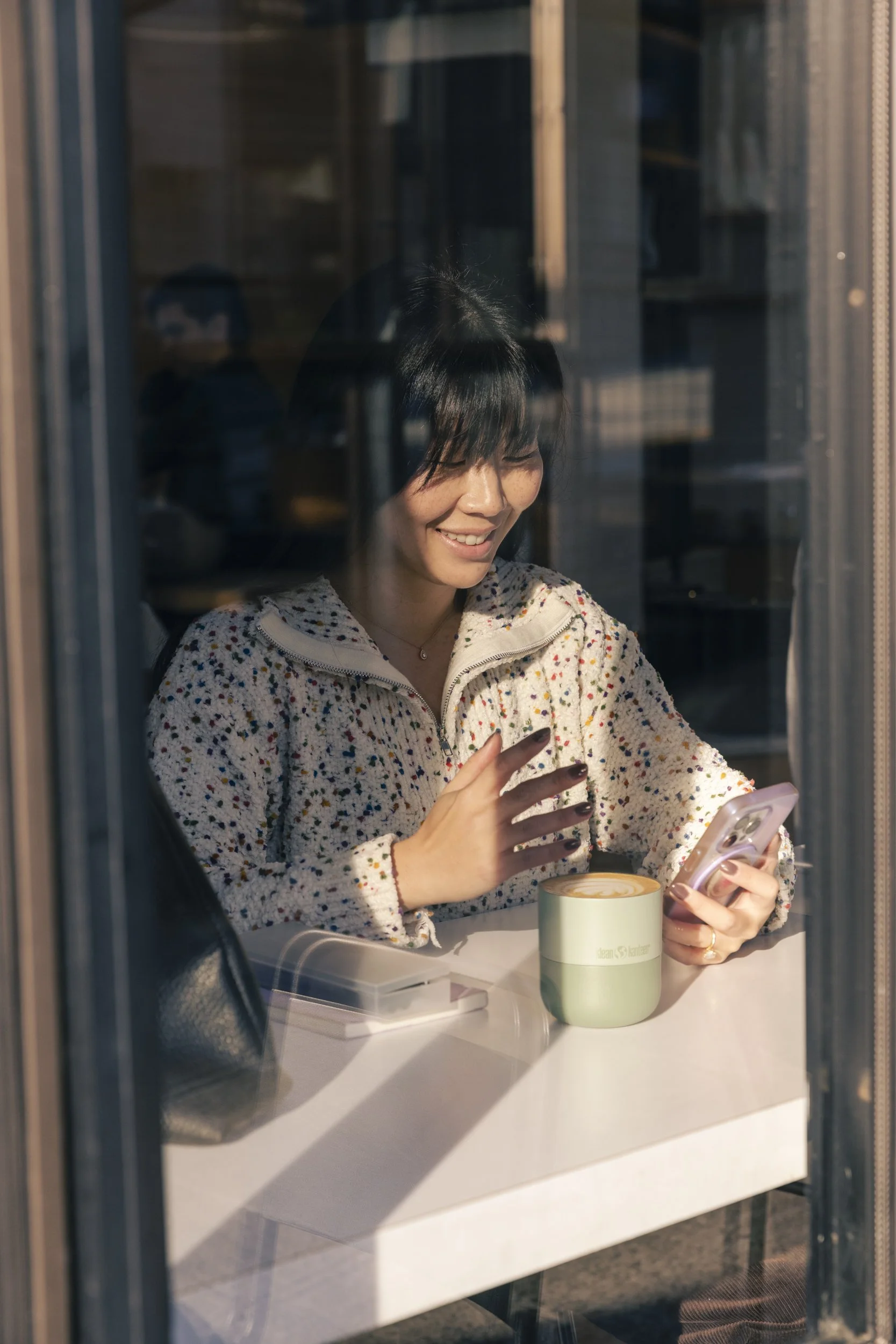 Product lifestyle photography by commercial photographer Kaare Iverson for Klean Kanteen.  Woman works from cafe in video meeting and reaches for reusable coffee mug - as seen through window.