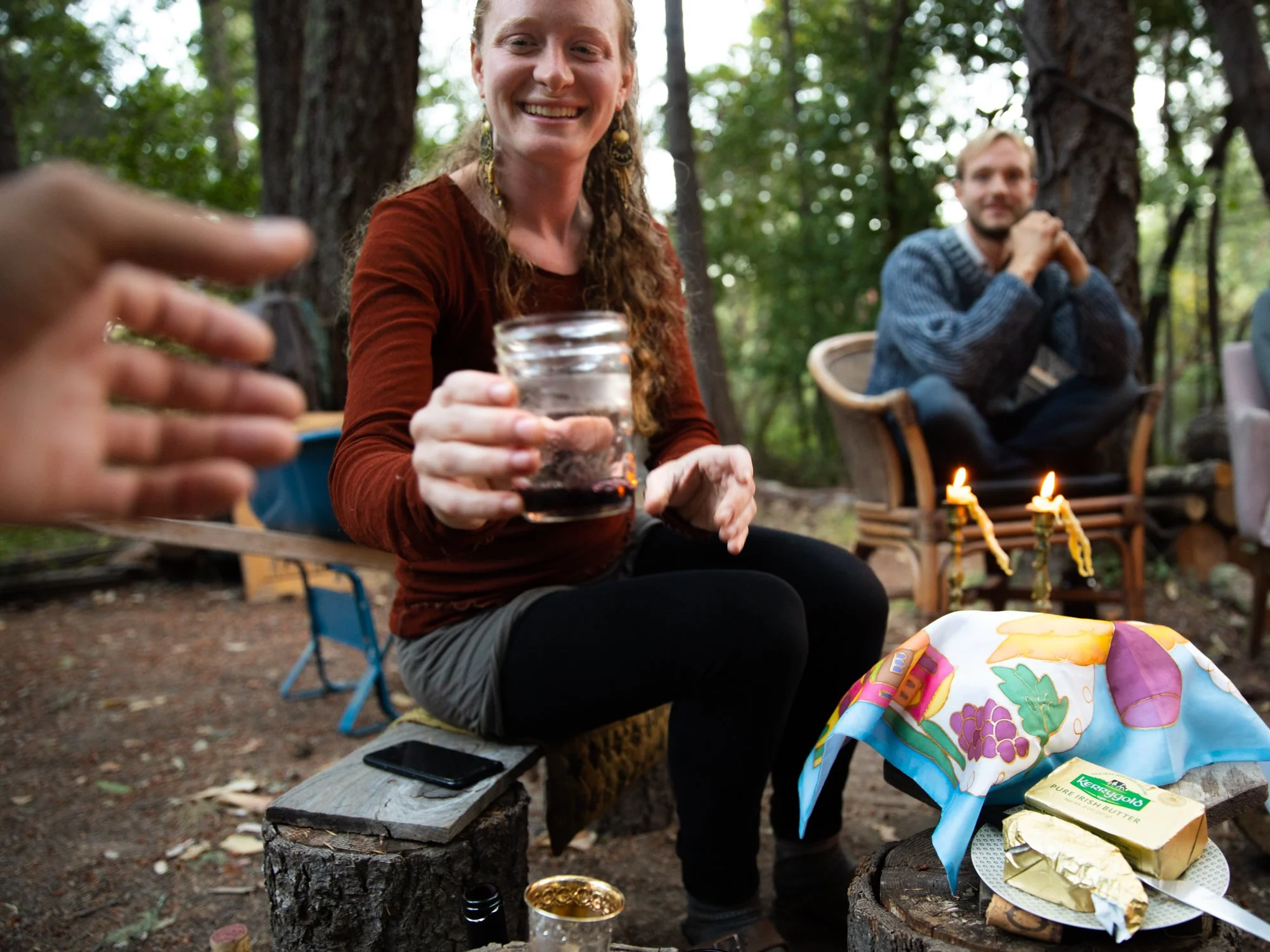 Image from PRAXIS, a photo essay shot in California by photographer Kaare Iverson. People are enjoying a gathering in a wooded outdoor setting. A woman in the foreground has long curly hair, earrings, and is holding a drink towards the camera.