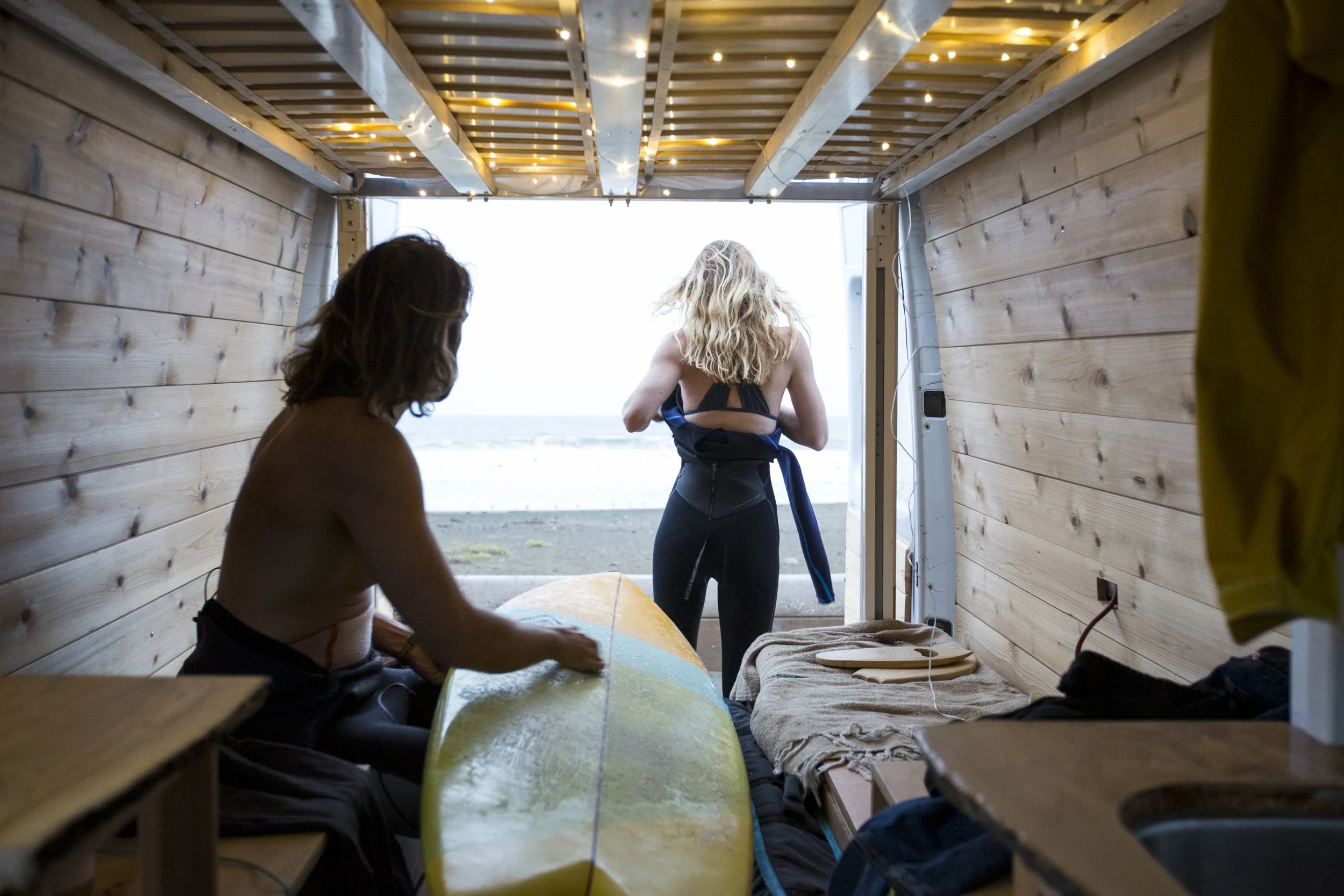 Outdoor adventure and travel lifestyle image of two surfers preparing their wetsuits inside a van overlooking the ocean in California's Bay Area by commercial photographer Kaare Iverson.