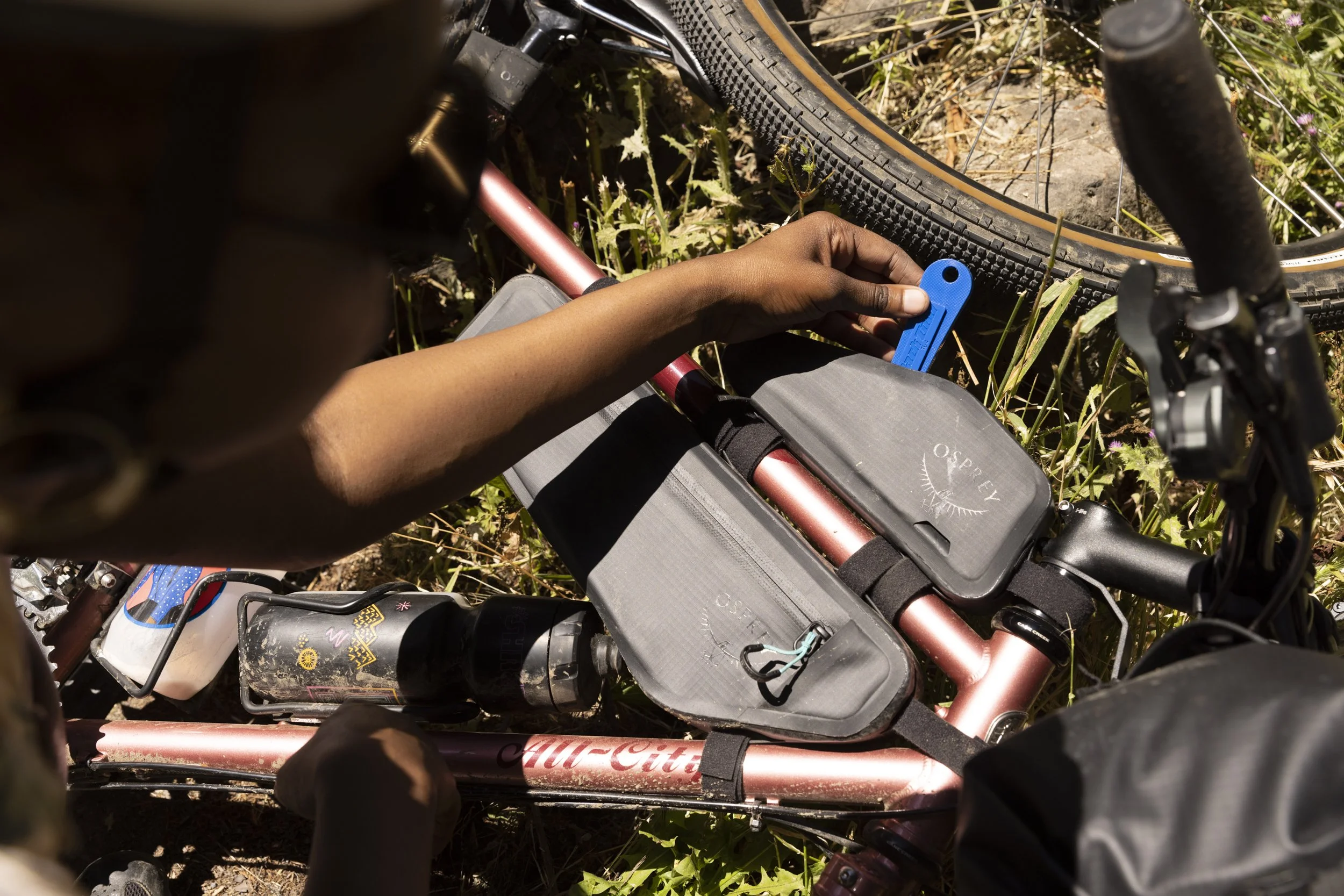 Product lifestyle photography of frame bag kit from Osprey Packs by California commercial photographer Kaare Iverson. Bicycle on the ground among grass, with a person's hand using a blue tool near the tire, possibly to repair or adjust the bike.