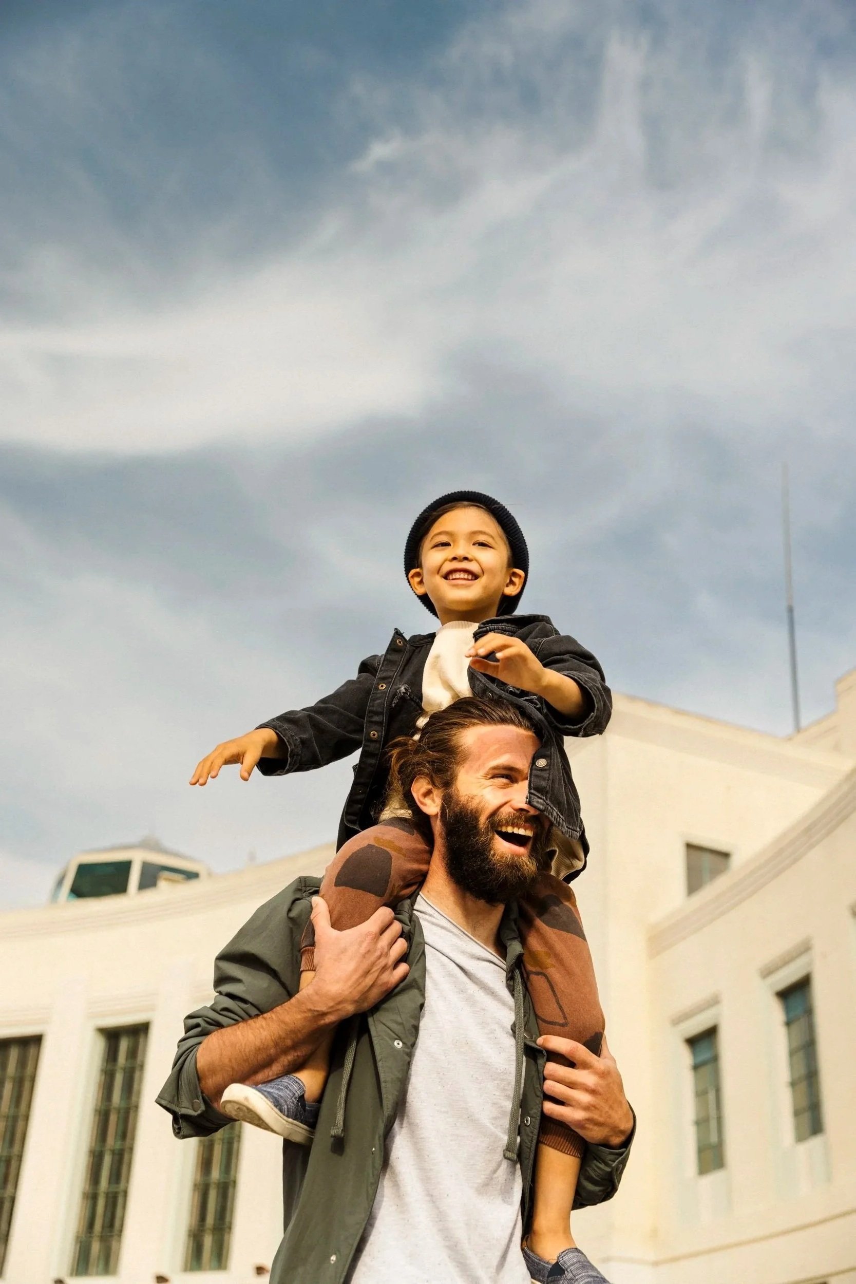 Commercial advertising photography for a real estate developer of a smiling man carrying a young boy on his shoulders outside in an urban area with modern buildings and a cloudy sky - shot in San Francisco, California.