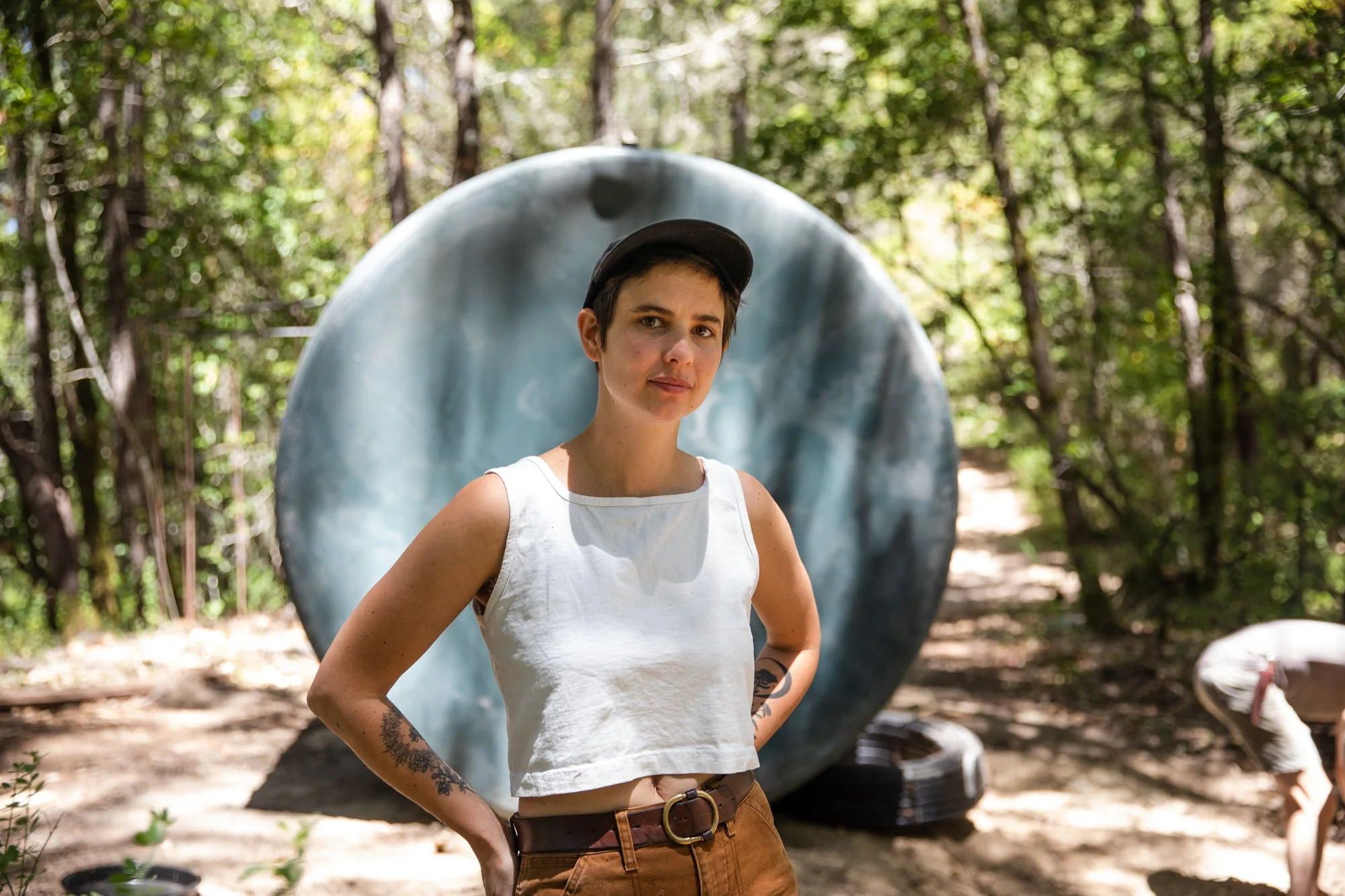 Image from PRAXIS, a photo essay shot in California by photographer Kaare Iverson. A woman with tattoos on her arms and short dark hair, wearing a black cap and a white tank top, standing outdoors in a forest with a water tank behind her.