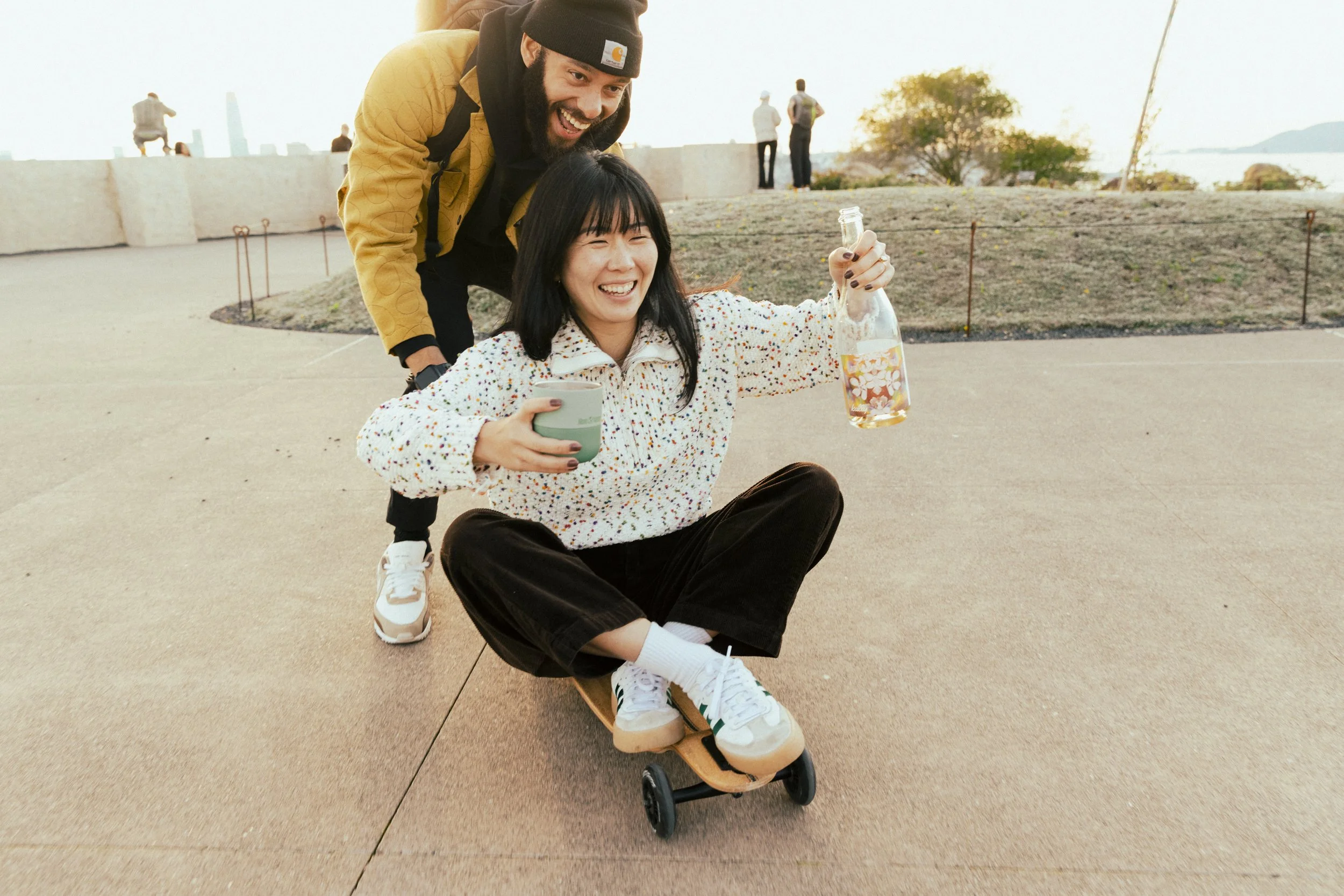 Urban playful lifestyle product photography by Bay Area based photographer Kaare Iverson.  Two friends push each other on skateboard while drinking Kally wine from a Klean Kanteen mug at sunset in an urban park in California.