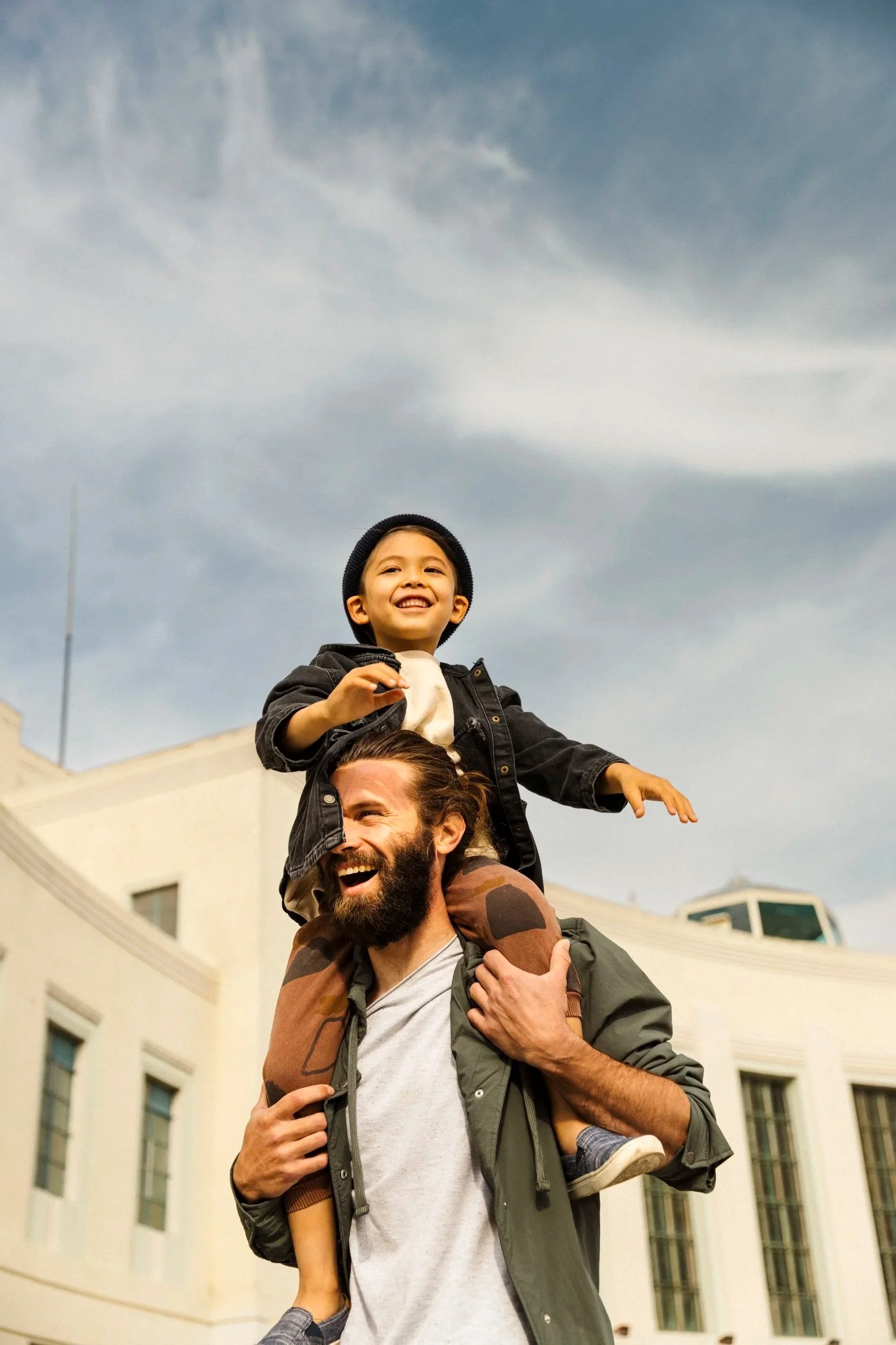 Lifestyle photography for San Francisco's Treasure Island Development by Kaare Iverson. A smiling man is carrying a young boy on his shoulders, who is also smiling. They are outdoors in front of a large, multi-story building with a cloudy sky above.