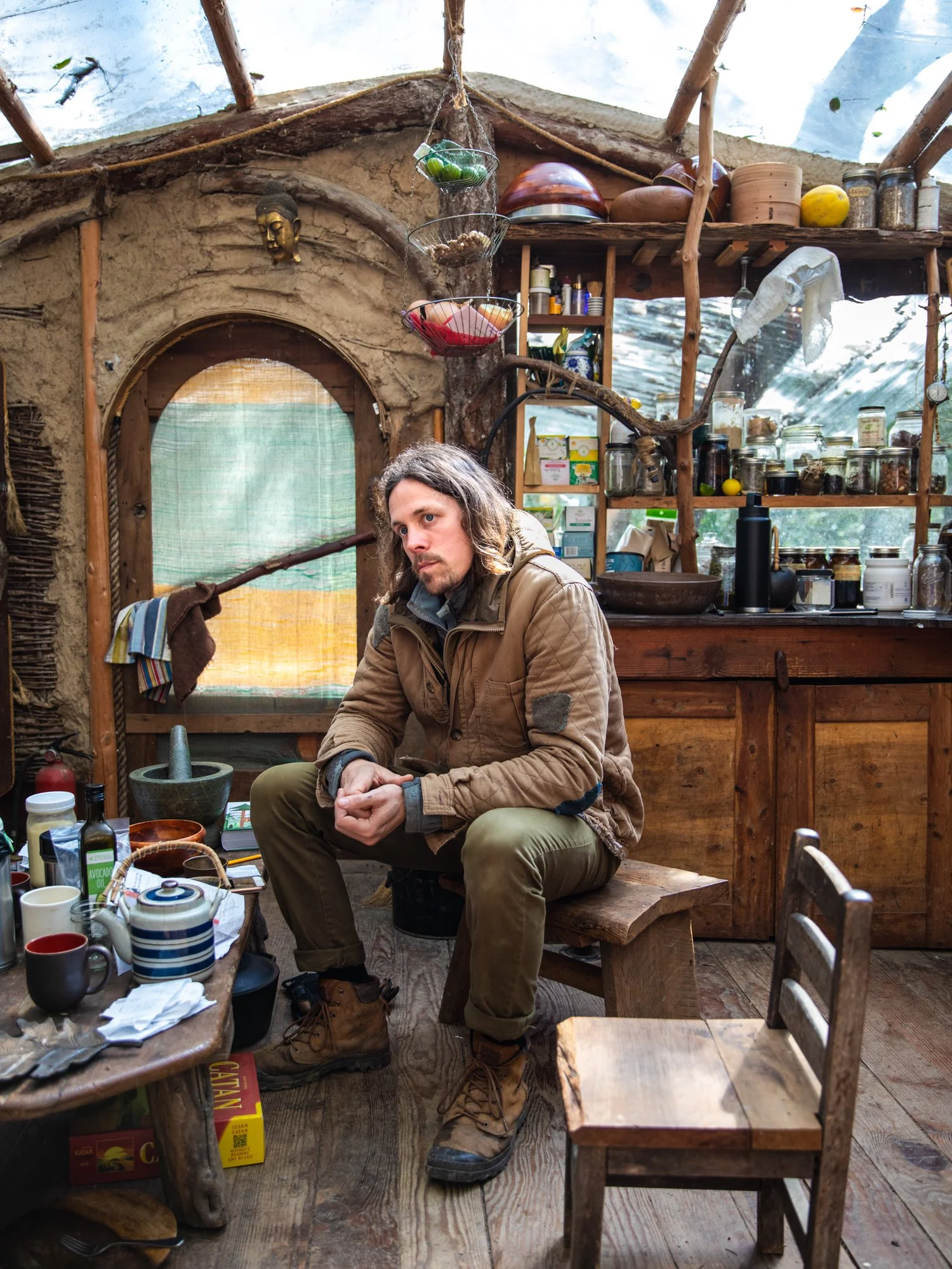 Image from the photo essay PRAXIS about alternative living in California by photographer Kaare Iverson. A man sitting on a wooden bench inside a rustic building surrounded by kitchen items, jars, and a window with a view of trees outside.