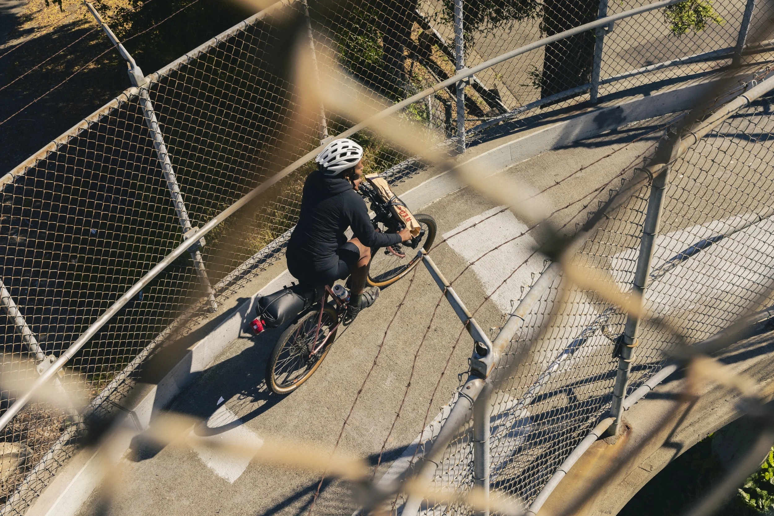 Cycling lifestyle photography for Osprey Packs frame bags.  A person riding a bicycle on a concrete pathway beneath a chain-link fence, taken from above through the fence.  Shot by commercial photographer Kaare Iverson in California.