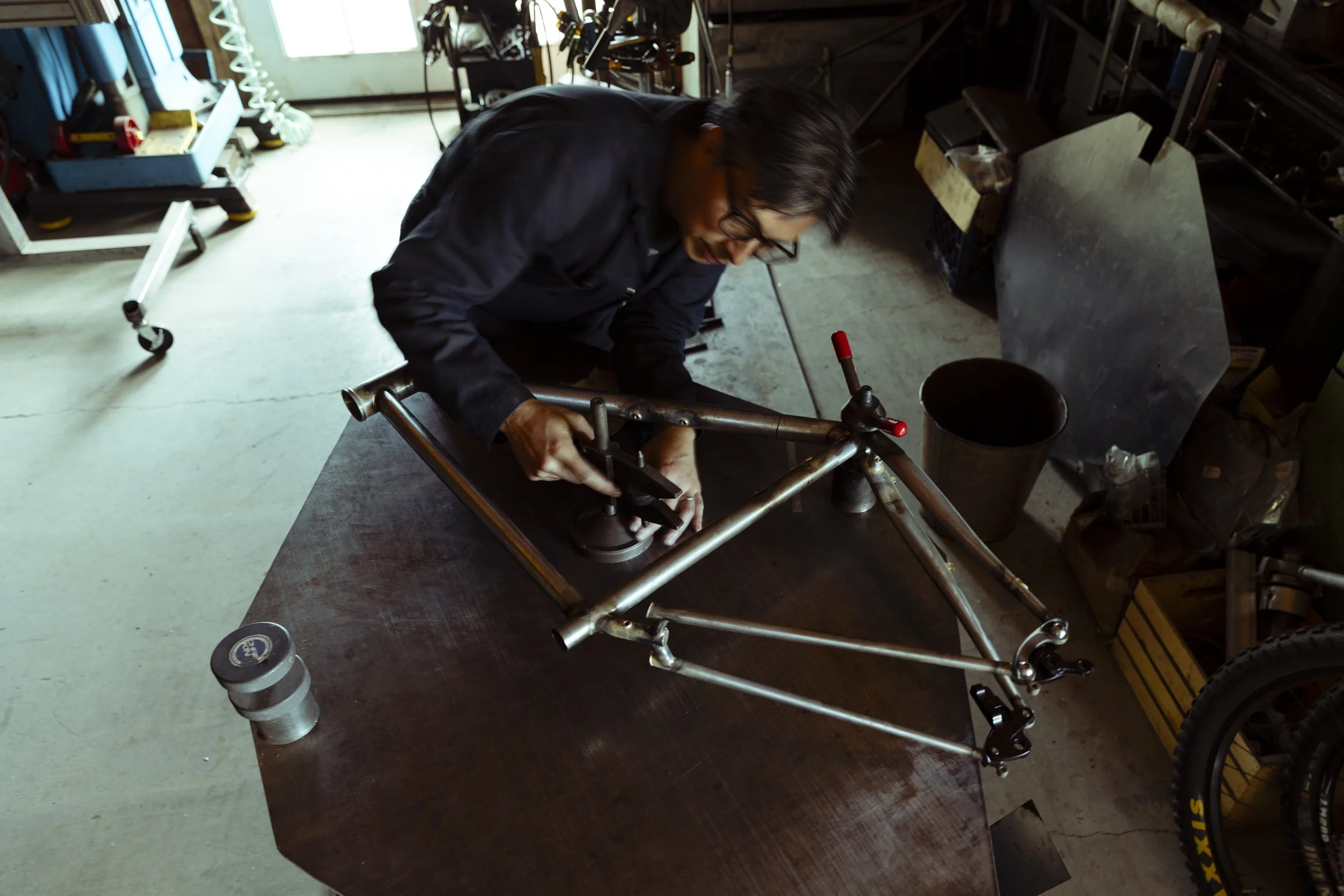 A man working on metal bike frame in workshop with tools and equipment surrounding him.