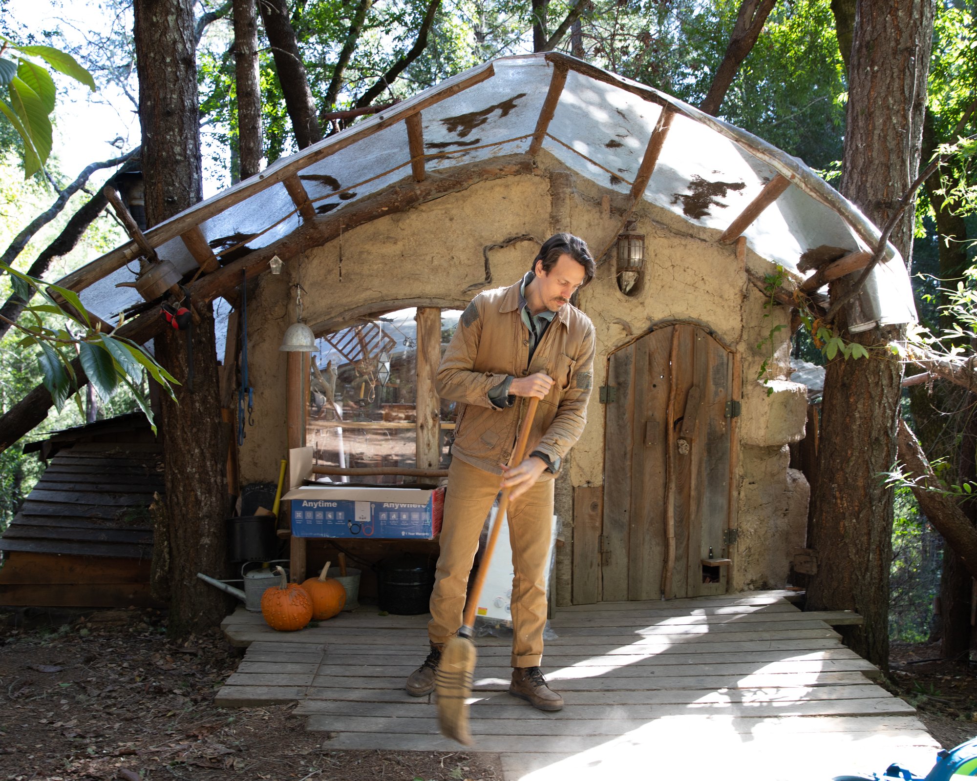 Image from PRAXIS, a photo essay shot in California by photographer Kaare Iverson. A man in outdoor work clothes sweeping a wooden deck in front of a rustic small house surrounded by trees. There are pumpkins on the ground near the house.