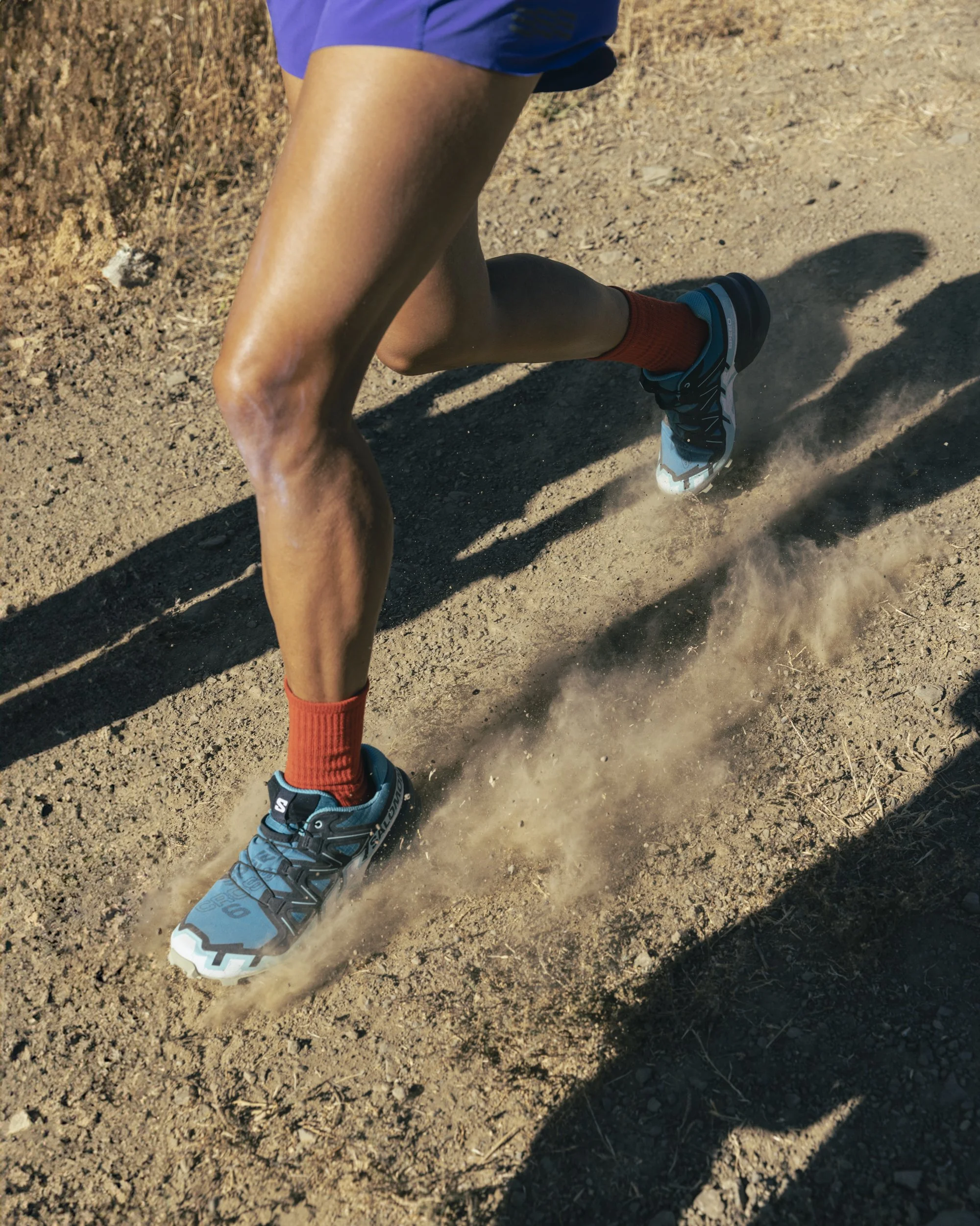 Athletic wear fitness lifestyle photography by San Francisco based photographer Kaare Iverson. A person running on a dirt trail wearing Salomon trailrunning shoes, red socks, and shorts, kicking up dust.