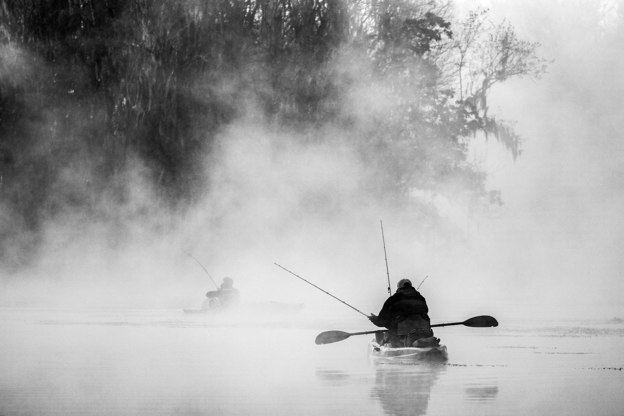 Two people are kayaking on a foggy body of water surrounded by trees, with mist rising from the water surface.