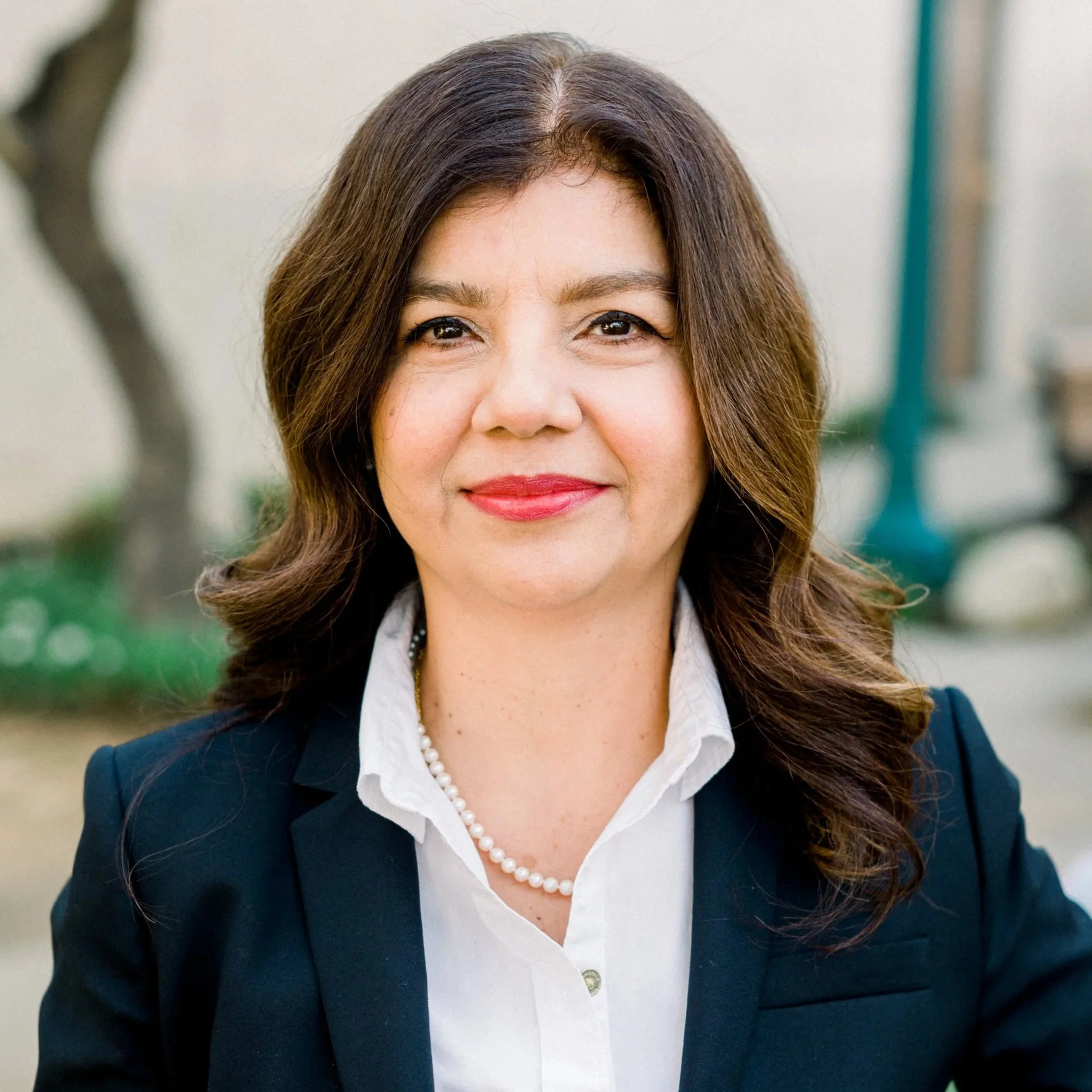 Los Angeles Lawyer Maria Mejia headshot wearing a navy blazer, white shirt, pearl necklace, and red lipstick, smiling outdoors.