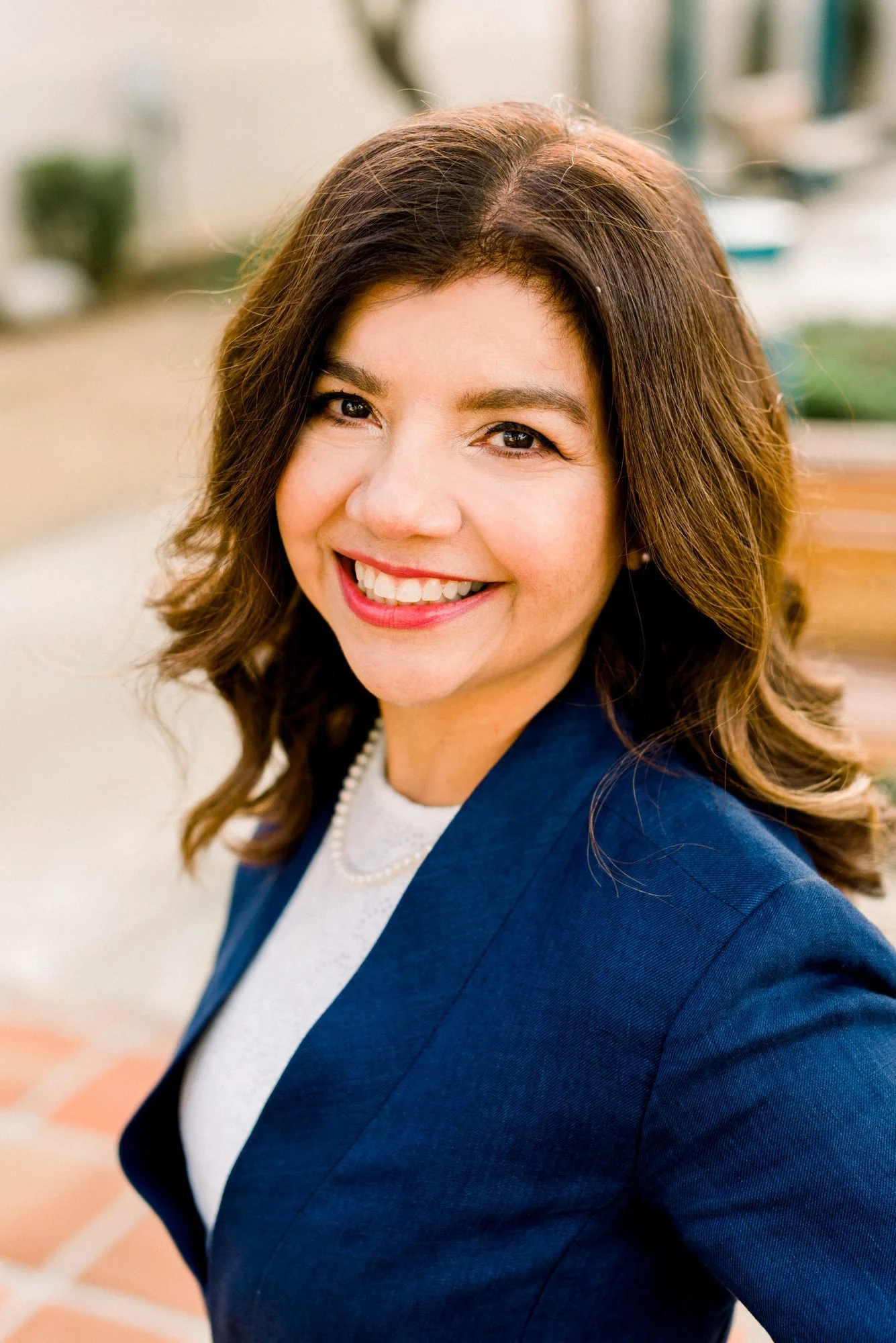 Family Law Attorney Maria Mejia with shoulder-length brown hair and a bright smile, wearing a navy blue blazer and white shirt, standing outdoors in front of a blurred background.