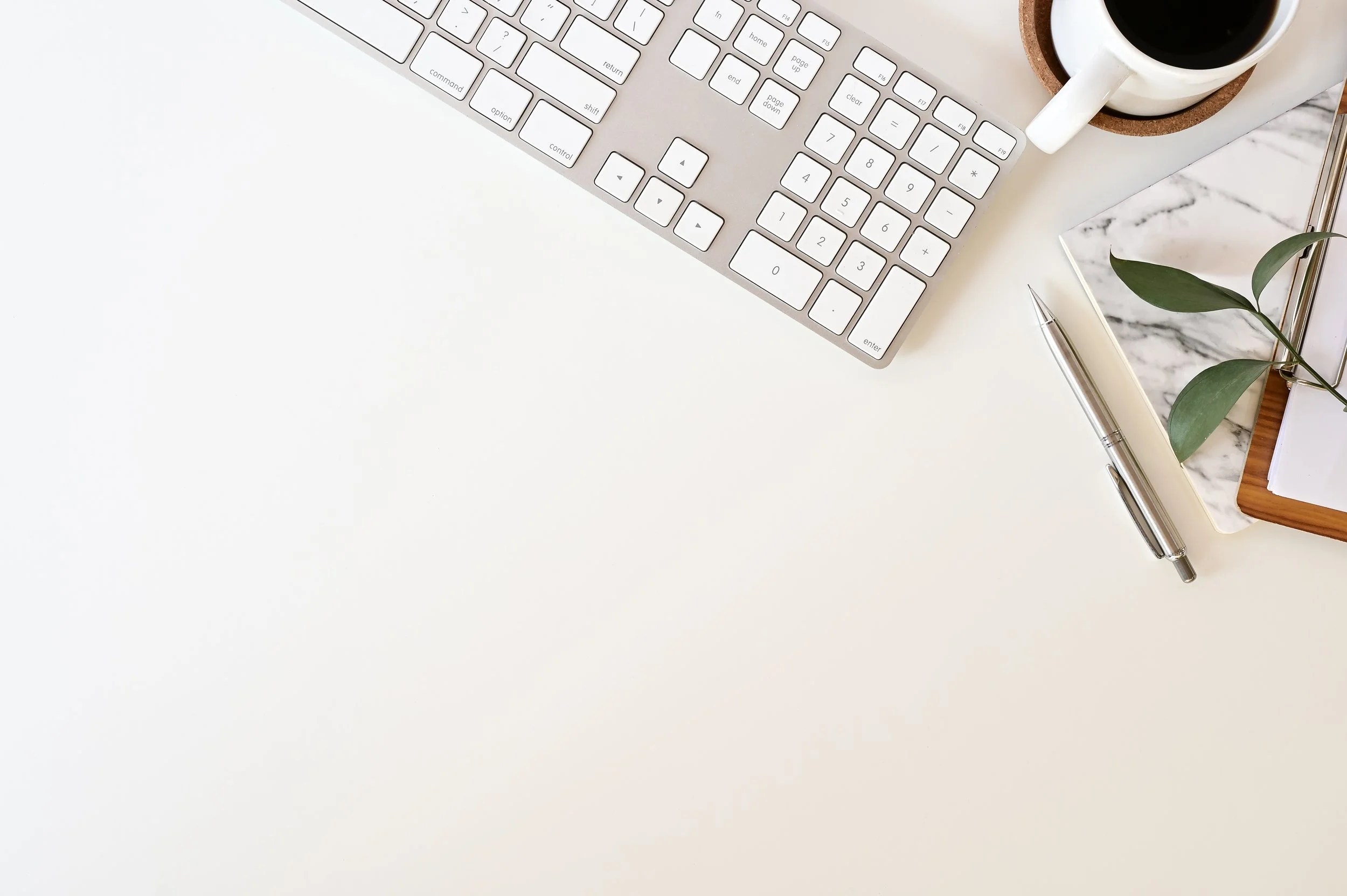 Top view of a computer keyboard, a cup of coffee, a notepad, a pen, and a green plant on a desk with a marble surface.