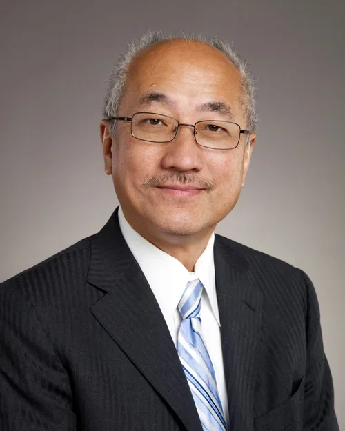 A middle-aged Asian man with short gray hair, glasses, and a slight smile, dressed in a black suit, white shirt, and a striped light blue tie, posed against a plain gray background.