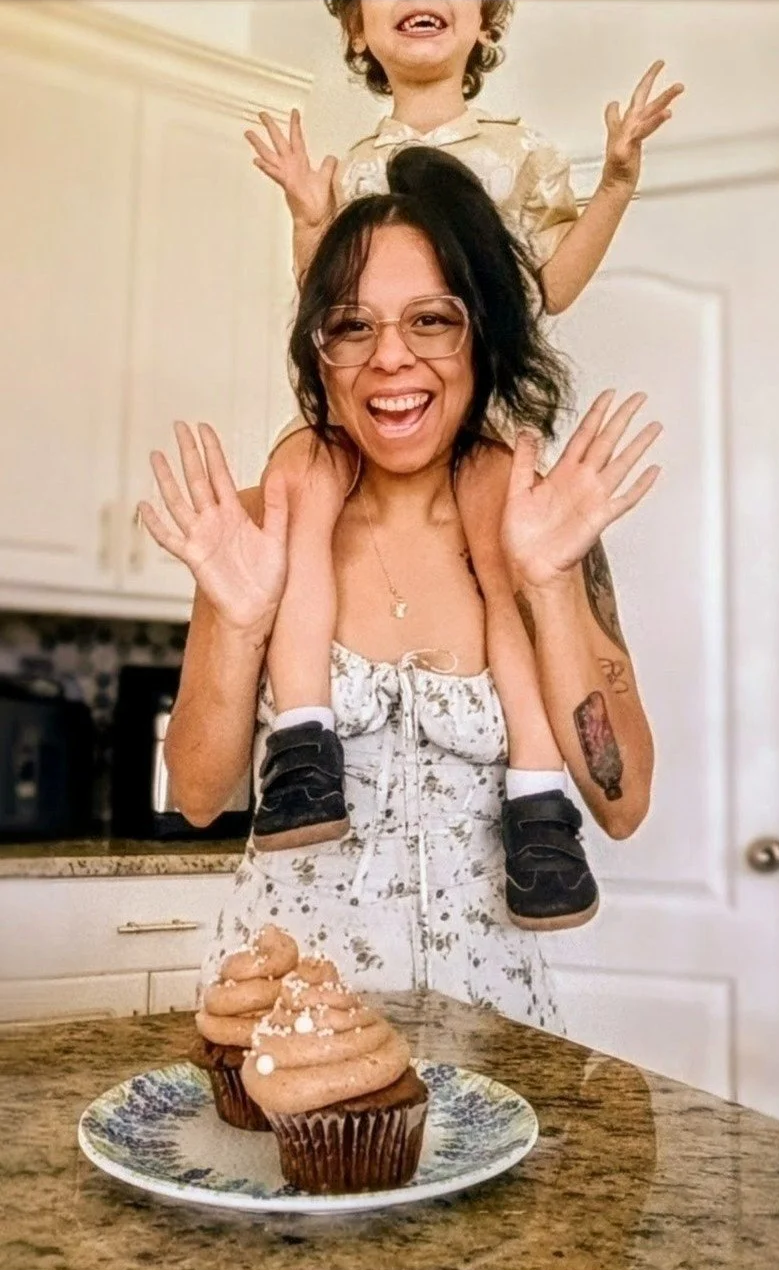 A woman with glasses and short dark hair is smiling and waving, carrying a young child on her shoulders. The child has curly hair and is also smiling. On the table in front of them are three cupcakes with swirled frosting and sprinkles.