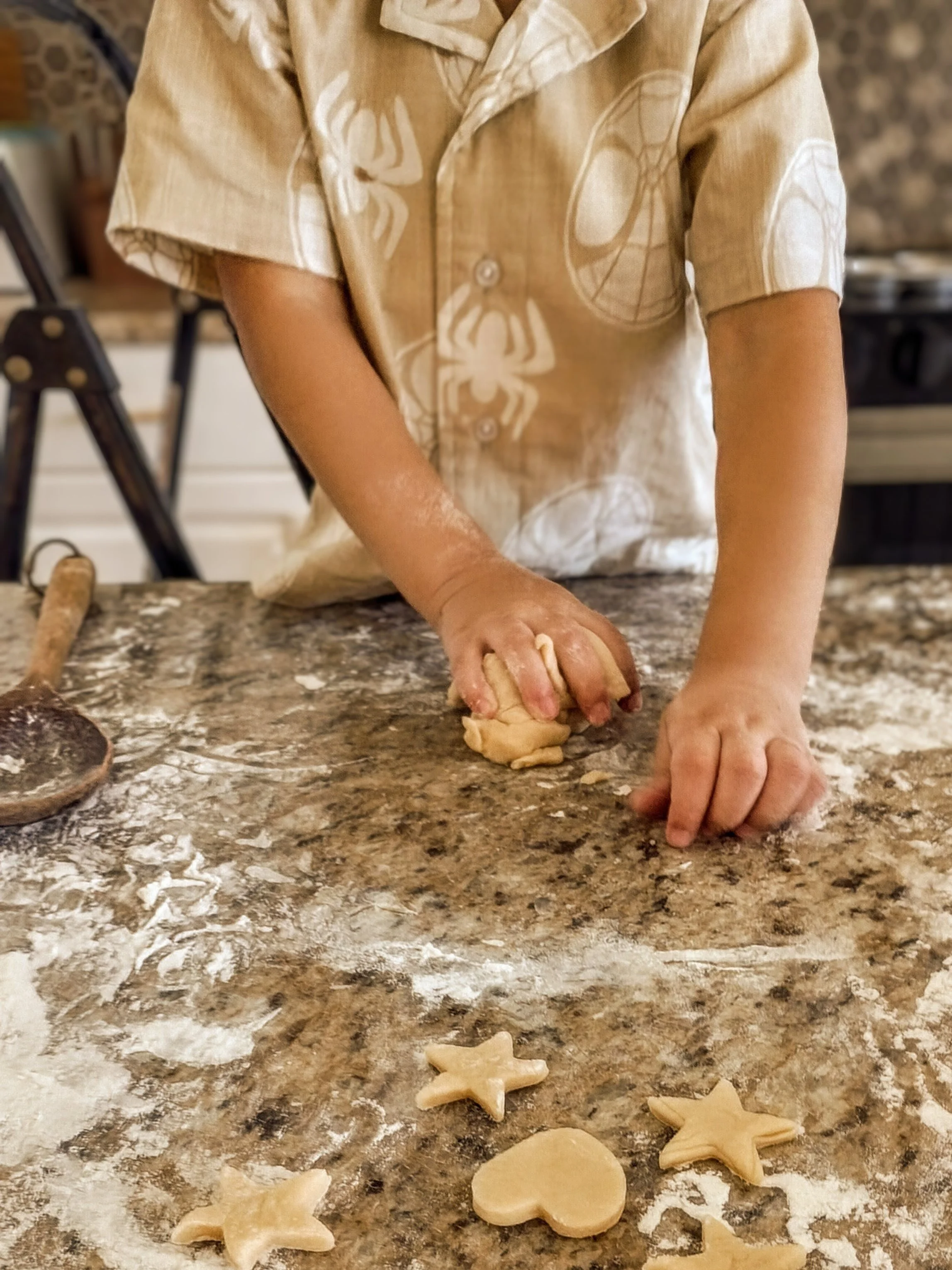 A person wearing a beige shirt with white spider and web patterns is kneading dough on a floured kitchen counter. Cookie cutters in star, heart, and circle shapes are on the counter, and a rolling pin is in the background.