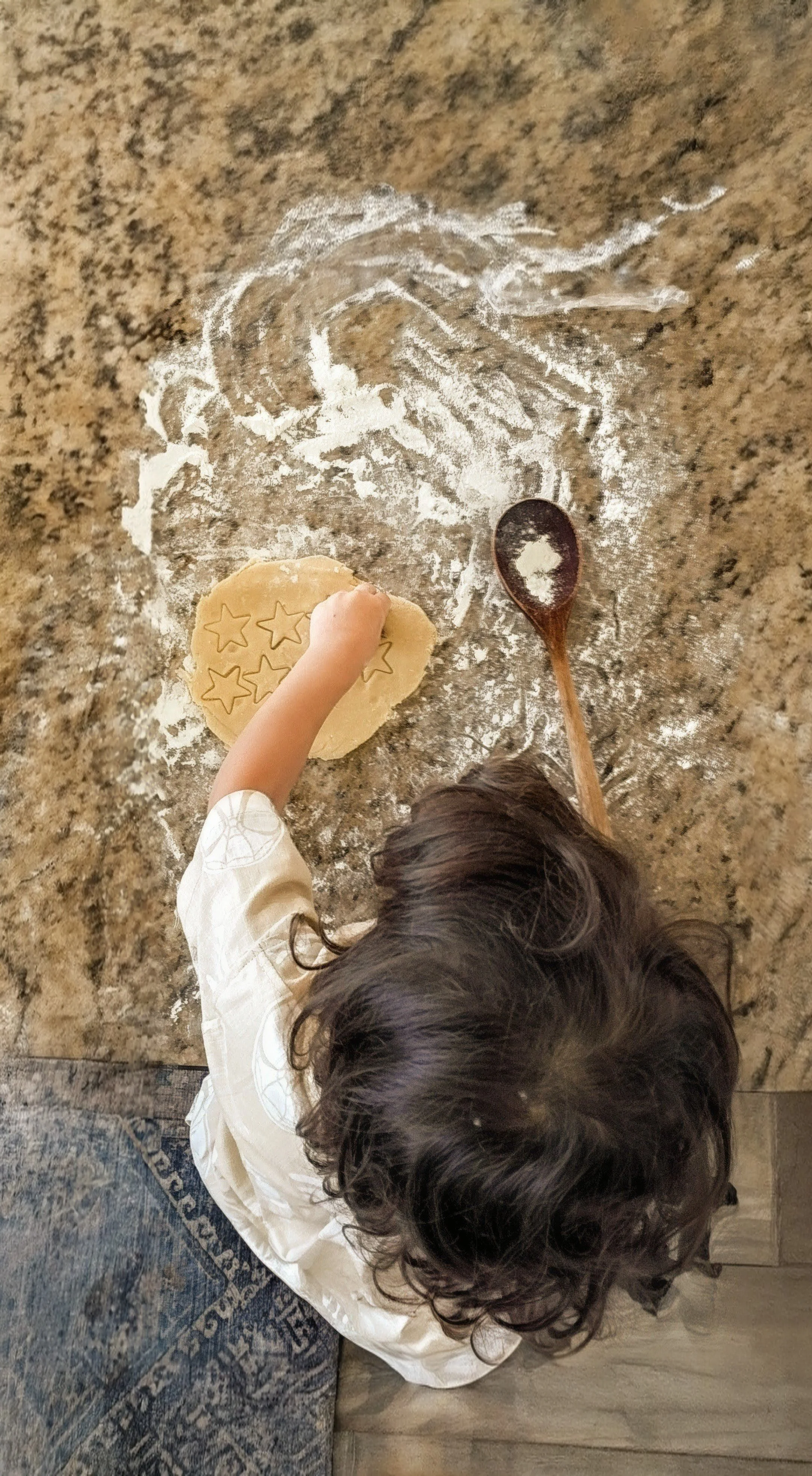 A child with long dark curly hair rolling out cookie dough with star-shaped cookie cutters on a granite countertop, dusted with flour, near a wooden spoon.