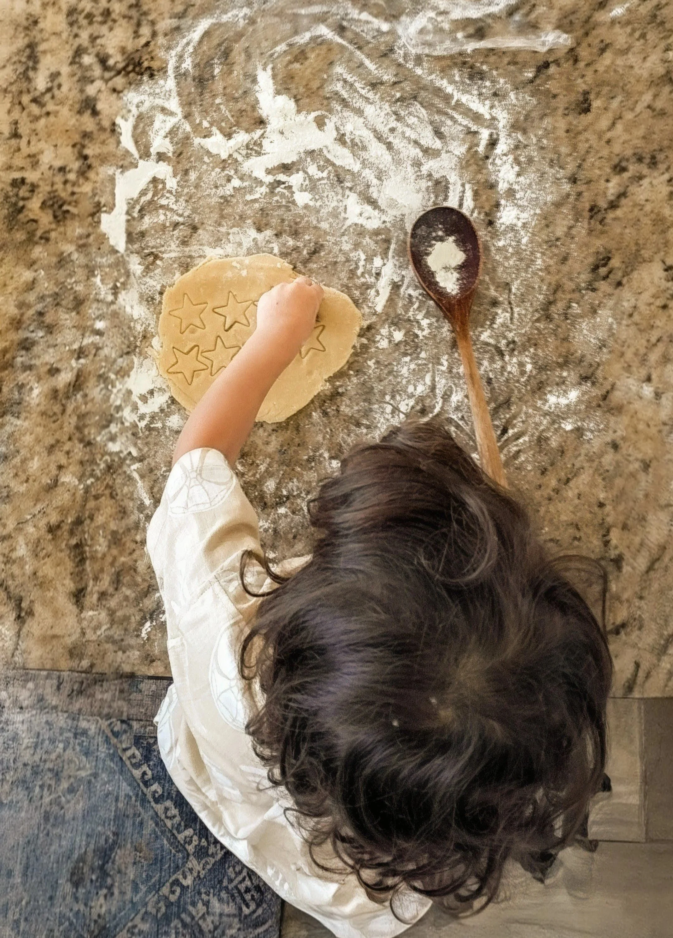 A child with dark curly hair wearing a white long-sleeve shirt, playing with star-shaped cookie cutters on a floured kitchen counter. A wooden spoon with flour on it is nearby.