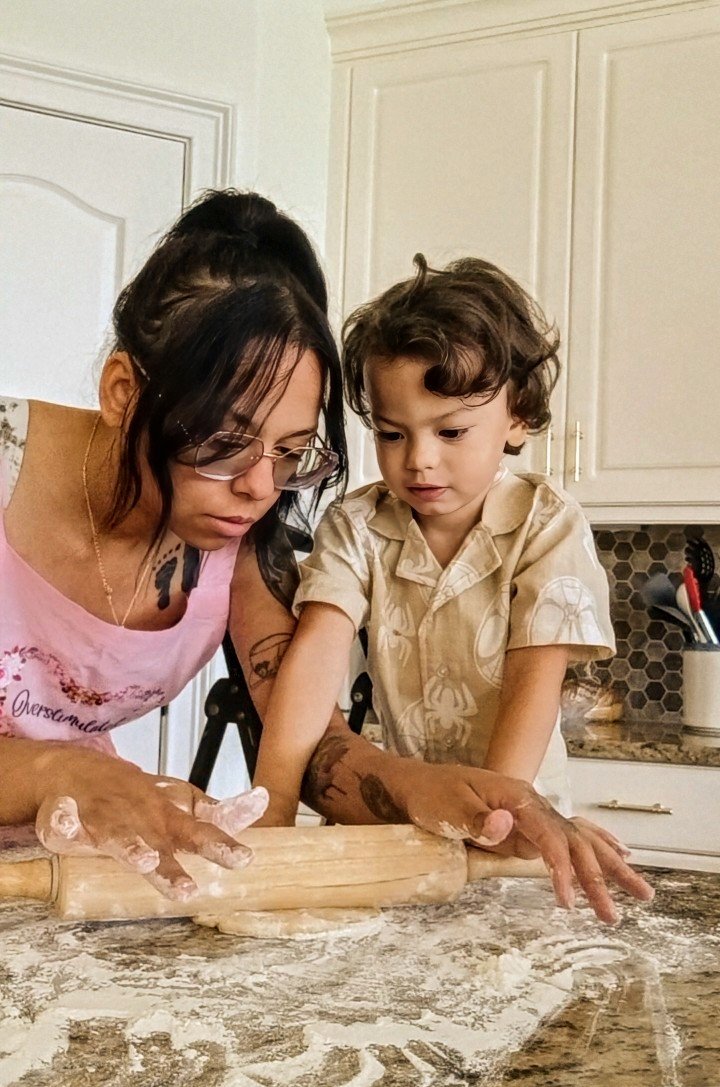 A woman and young boy baking together in a kitchen. They are using a rolling pin on dough on a marble countertop, with flour sprinkled around.
