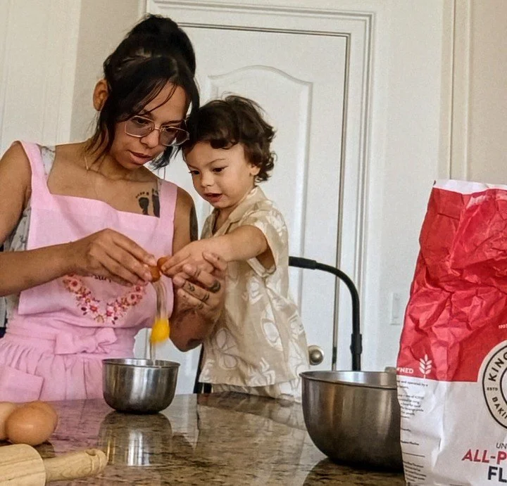 A woman and a young boy are cracking an egg into a metal bowl on a kitchen counter. The woman is wearing glasses and a pink apron, and the boy is wearing a cream-colored shirt. There is a bag of flour, another bowl, and a few eggs on the counter.