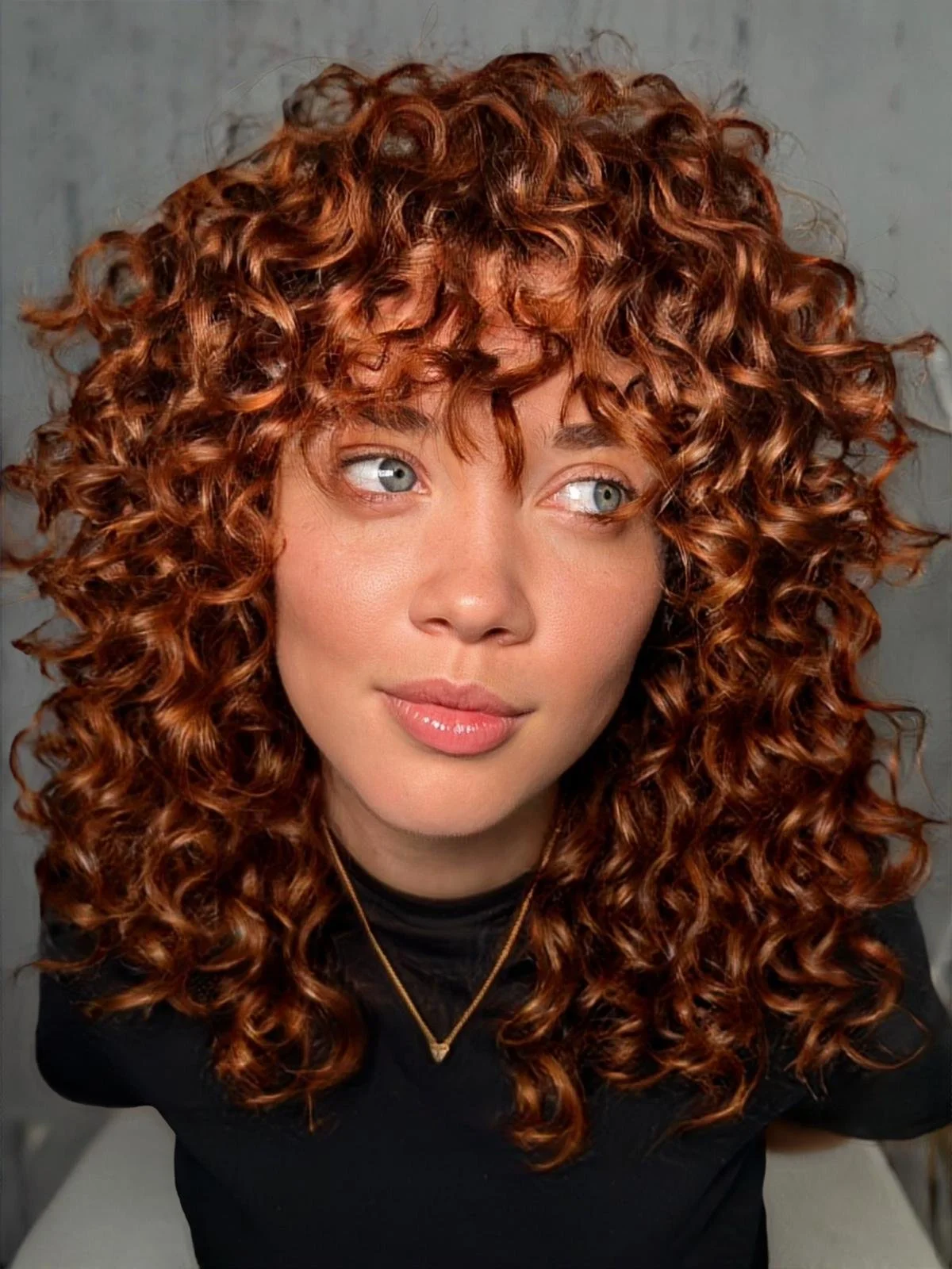 Close-up portrait of a woman with voluminous, curly red hair, wearing a black top and a delicate gold necklace, looking thoughtfully to her left.