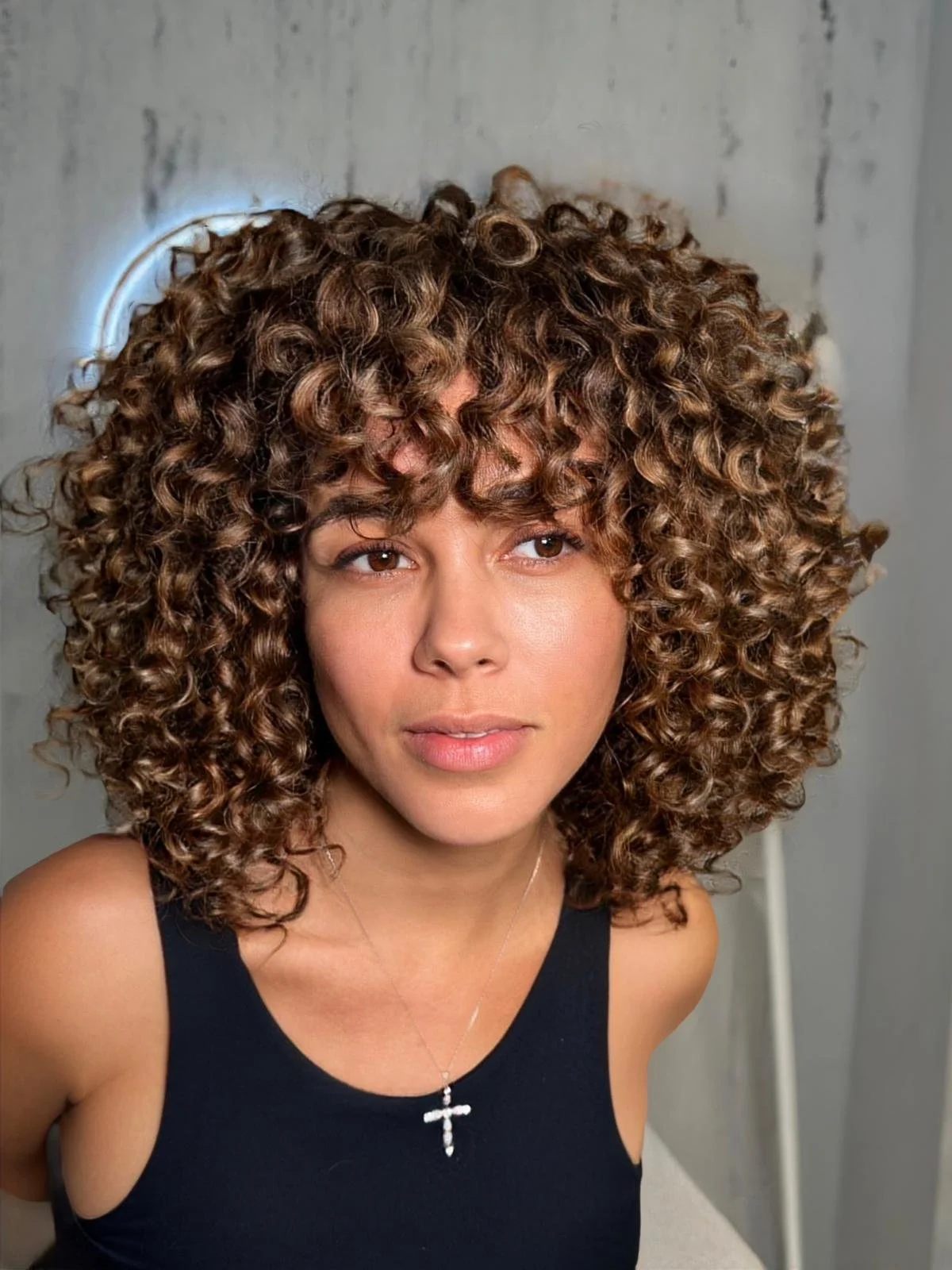 A woman with tightly coiled, brown hair styled in a voluminous afro, wearing a black tank top and a silver cross necklace, looking slightly to her right, with a neutral background.
