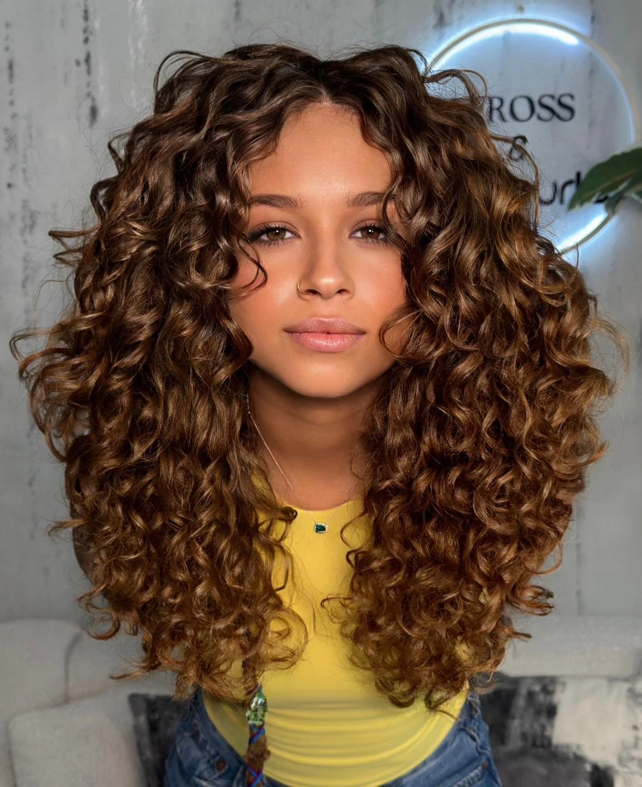 A woman with long, voluminous curly brown hair, wearing a yellow top and a necklace, posing in front of a wall with a round illuminated sign.
