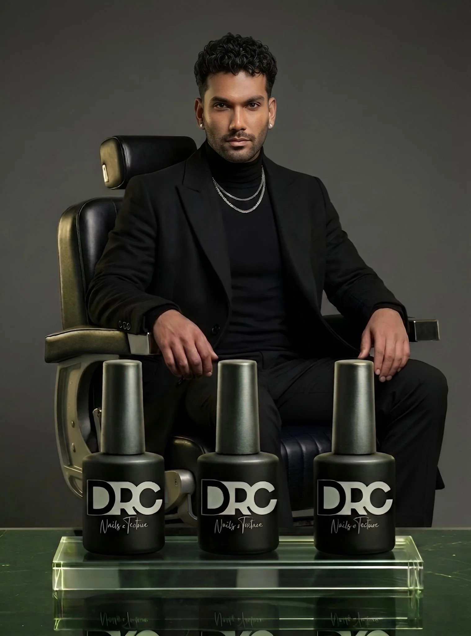 A man with dark, curly hair and earrings, dressed in black, seated in a black chair against a gray background, with three black containers labeled 'DRC' with white text and a signature, placed in front of him on a clear surface.