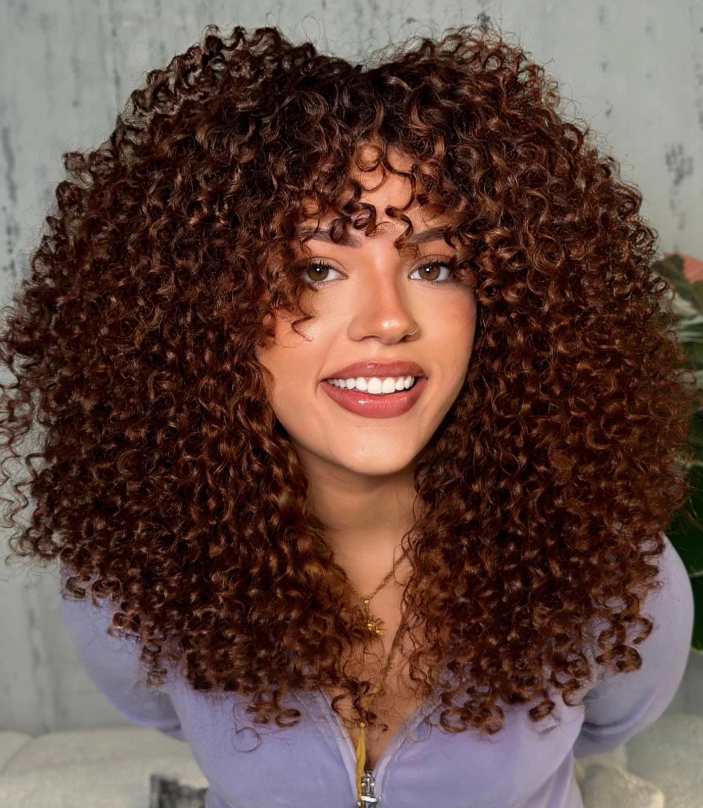 A woman with curly, voluminous, auburn hair smiling at the camera, wearing a light purple zip-up top, gold jewelry, and makeup.
