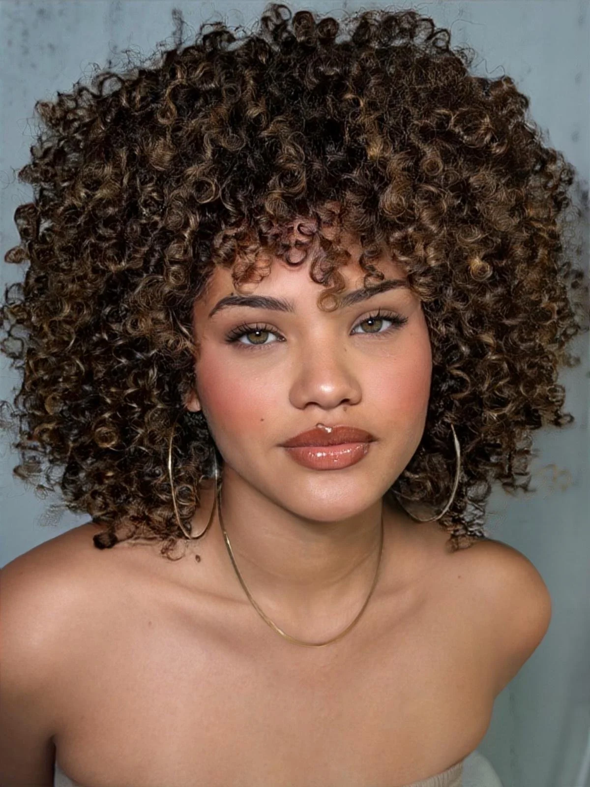 A woman with curly brown hair, hazel eyes, and a radiant smile. She is wearing hoop earrings, a gold necklace, and makeup with glossy lips and blush. The background is plain and light-colored.