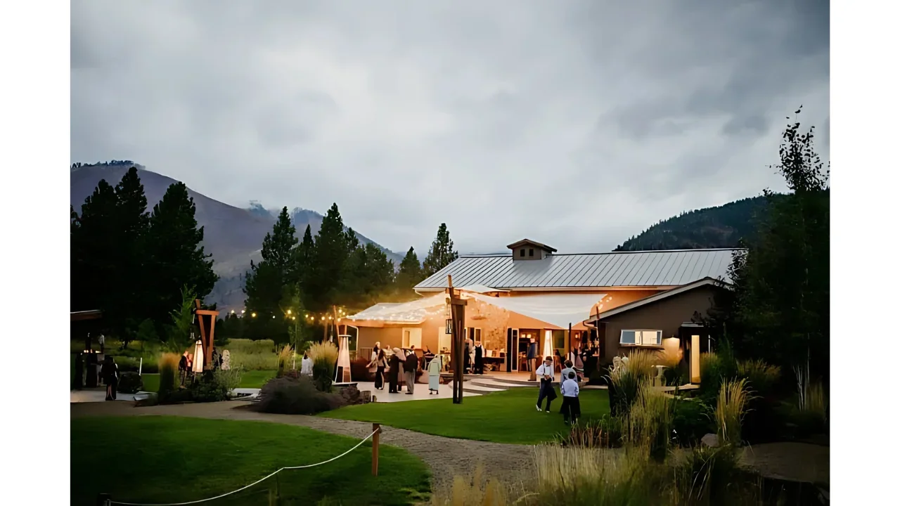 People socializing at an outdoor evening event with string lights, in front of a modern building surrounded by greenery and mountains under a cloudy sky.